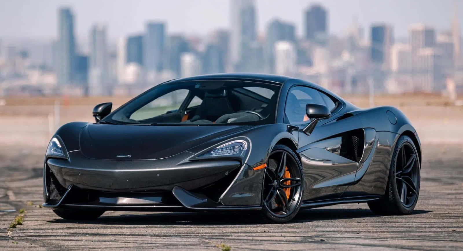 Mclaren Gt 桌布 壁紙 — 一輛時尚的黑色McLaren跑車停在混凝土場地上，背景是模糊的城市天際線。