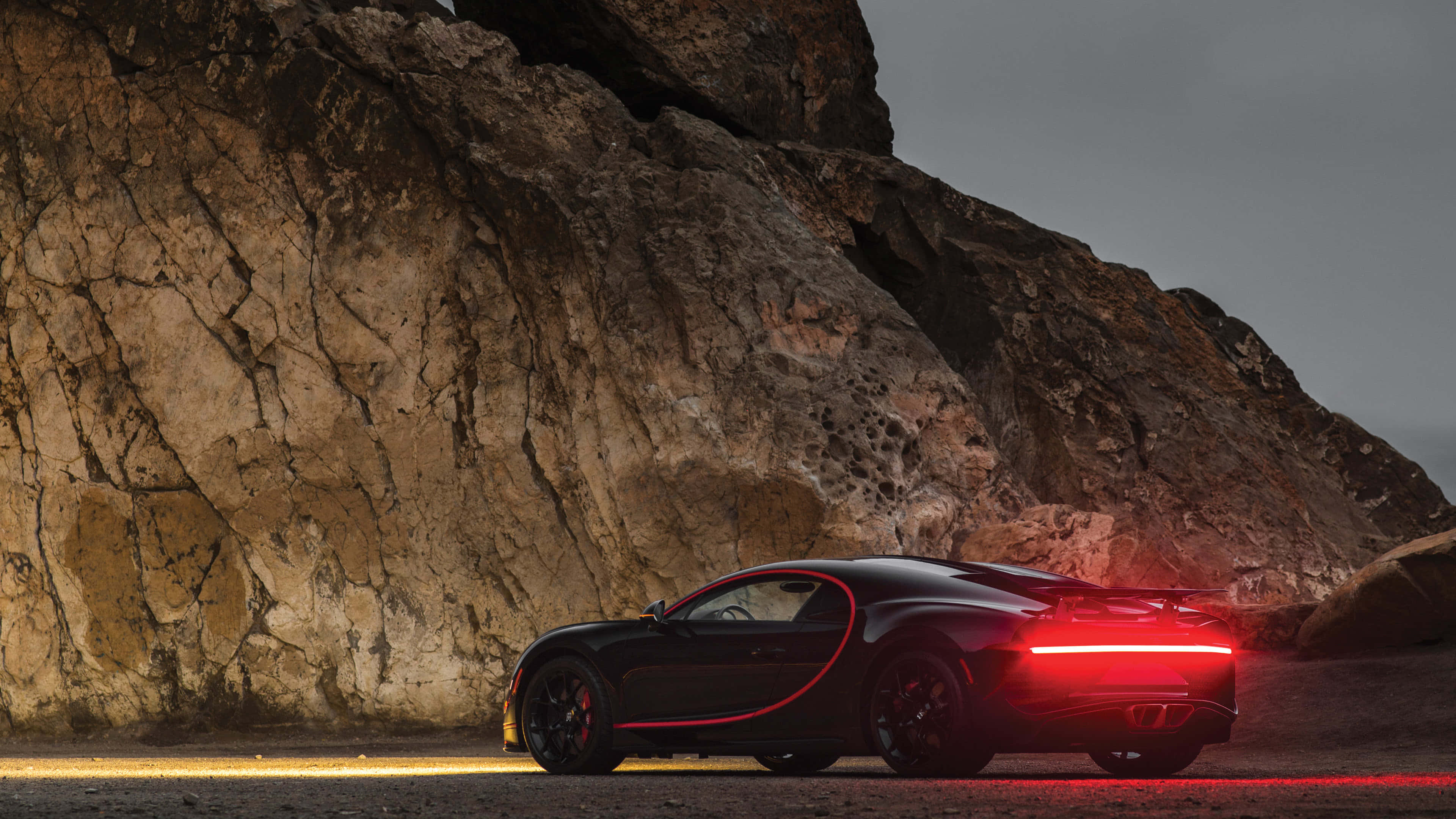 Bugatti Chiron 桌布 壁紙 — 一輛時尚的黑色跑車，帶有發光的紅色尾燈，停泊在黃昏時崎嶇的岩石懸崖旁。