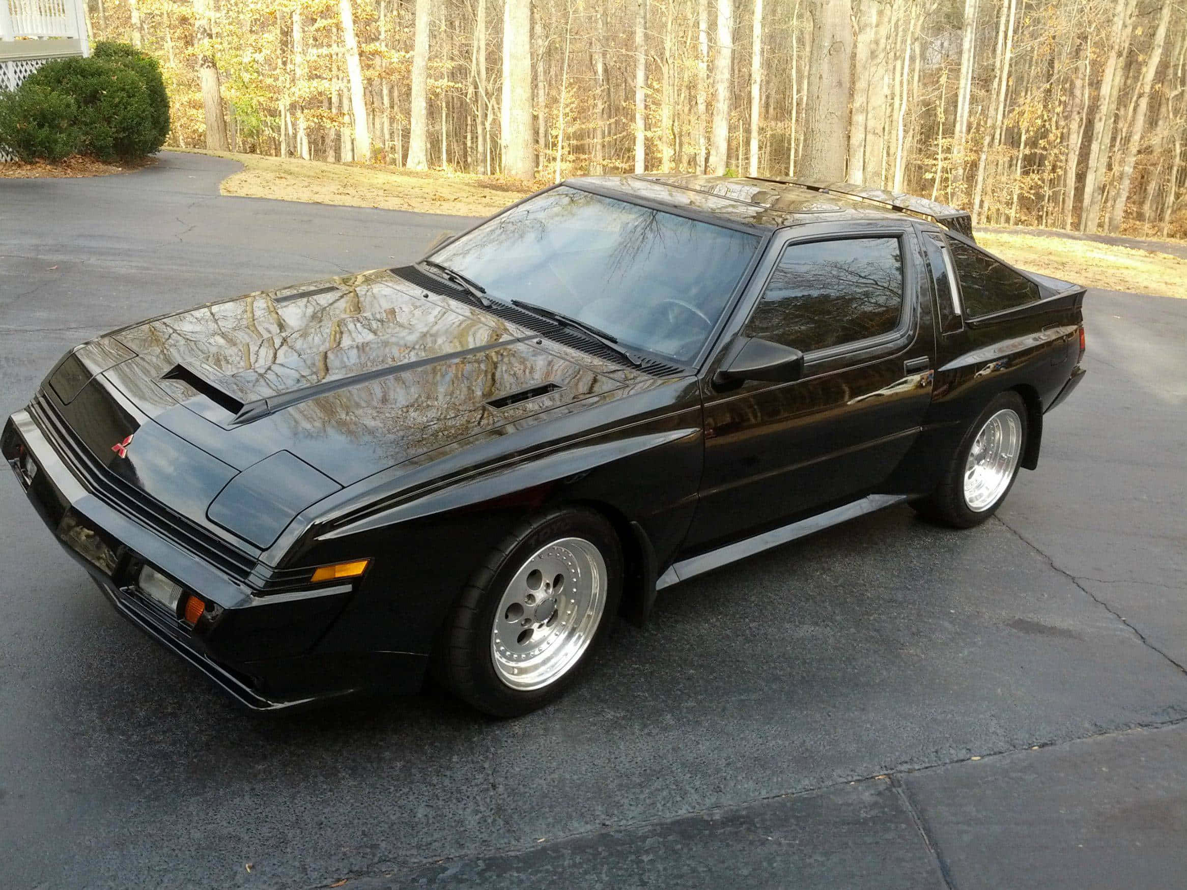Mitsubishi Starion 桌布 壁紙 — 一輛時尚的黑色三菱Starion跑車停放在車道上，光亮的引擎蓋反射出光禿禿的樹木。
