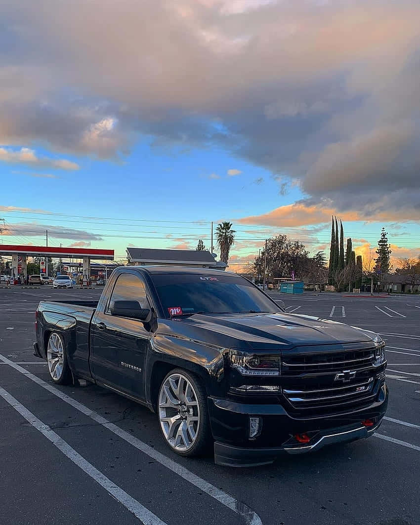  壁紙 — 一輛時尚的黑色雪佛蘭 Silverado 皮卡車，配備定制輪圈，停泊在加油站內，背景是戲劇性的日落天空。