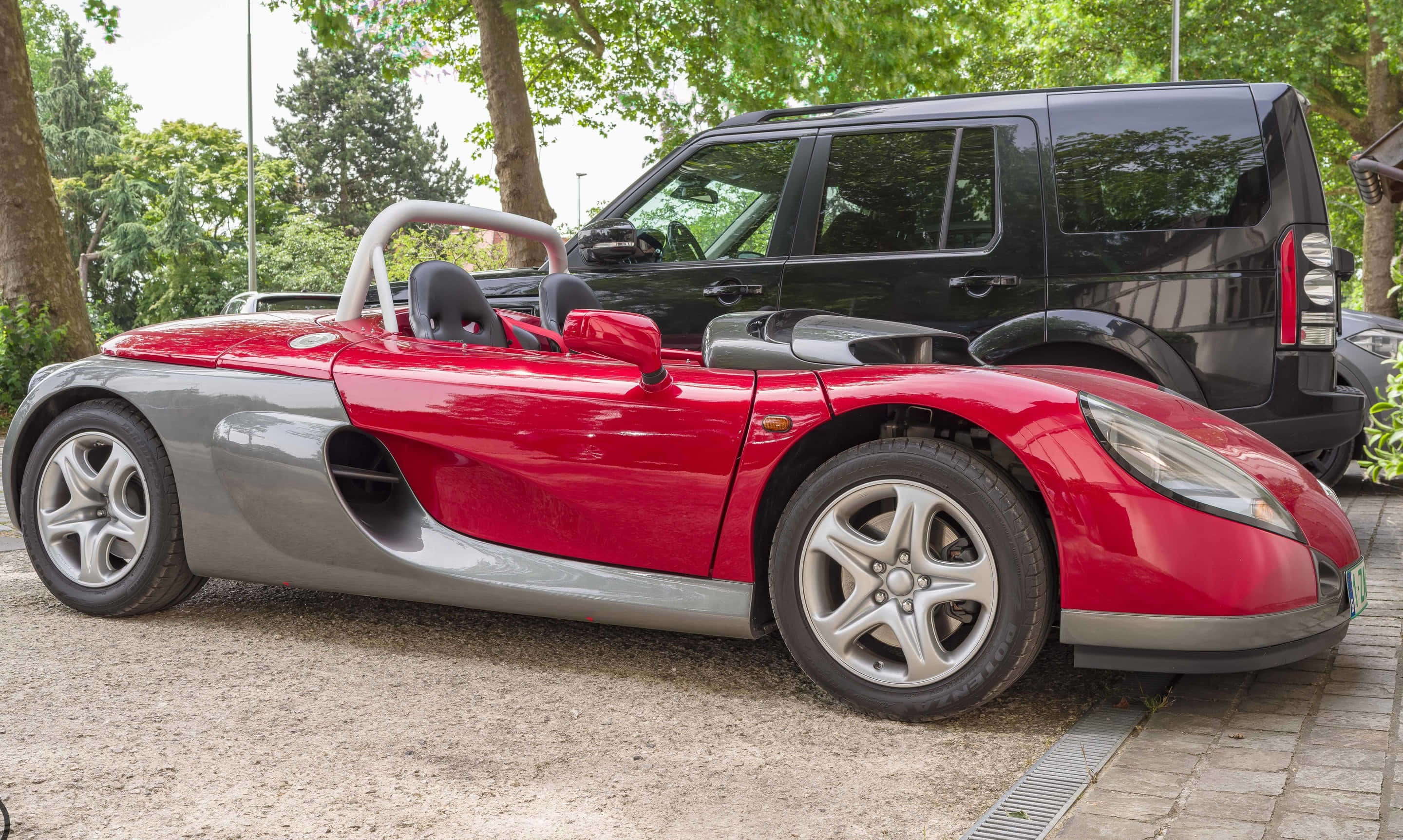 Renault Sport Spider 桌布 壁紙 — 一輛時尚的紅色和銀色跑車，配有防滾籠，停在一輛黑色SUV旁邊，位於樹葉茂密的樹下。
