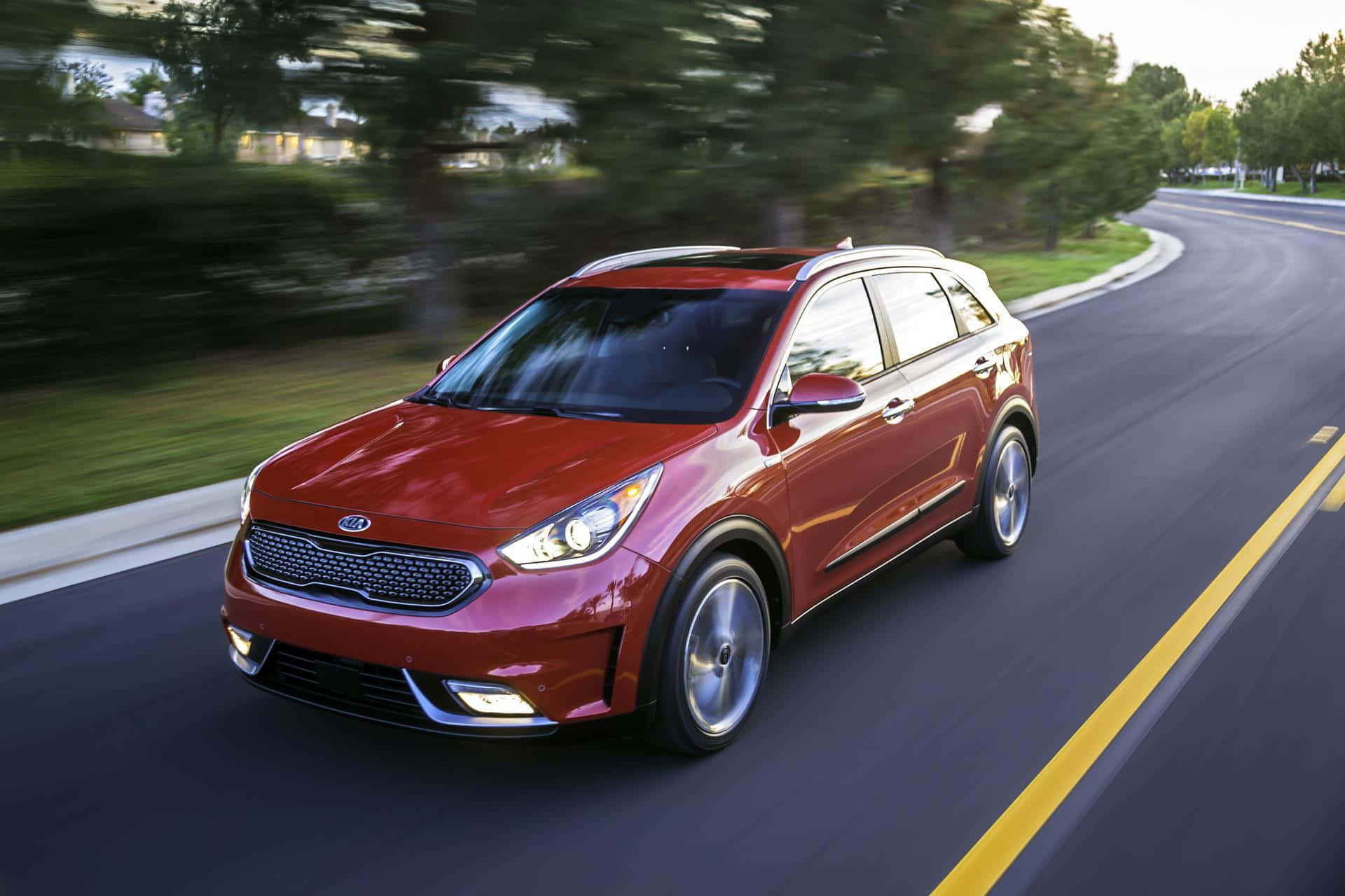 Kia Niro 桌布 壁紙 — 一輛時尚的紅色Kia SUV在彎曲的郊區道路上飛馳，背景是模糊的綠樹和金色的日落光線。