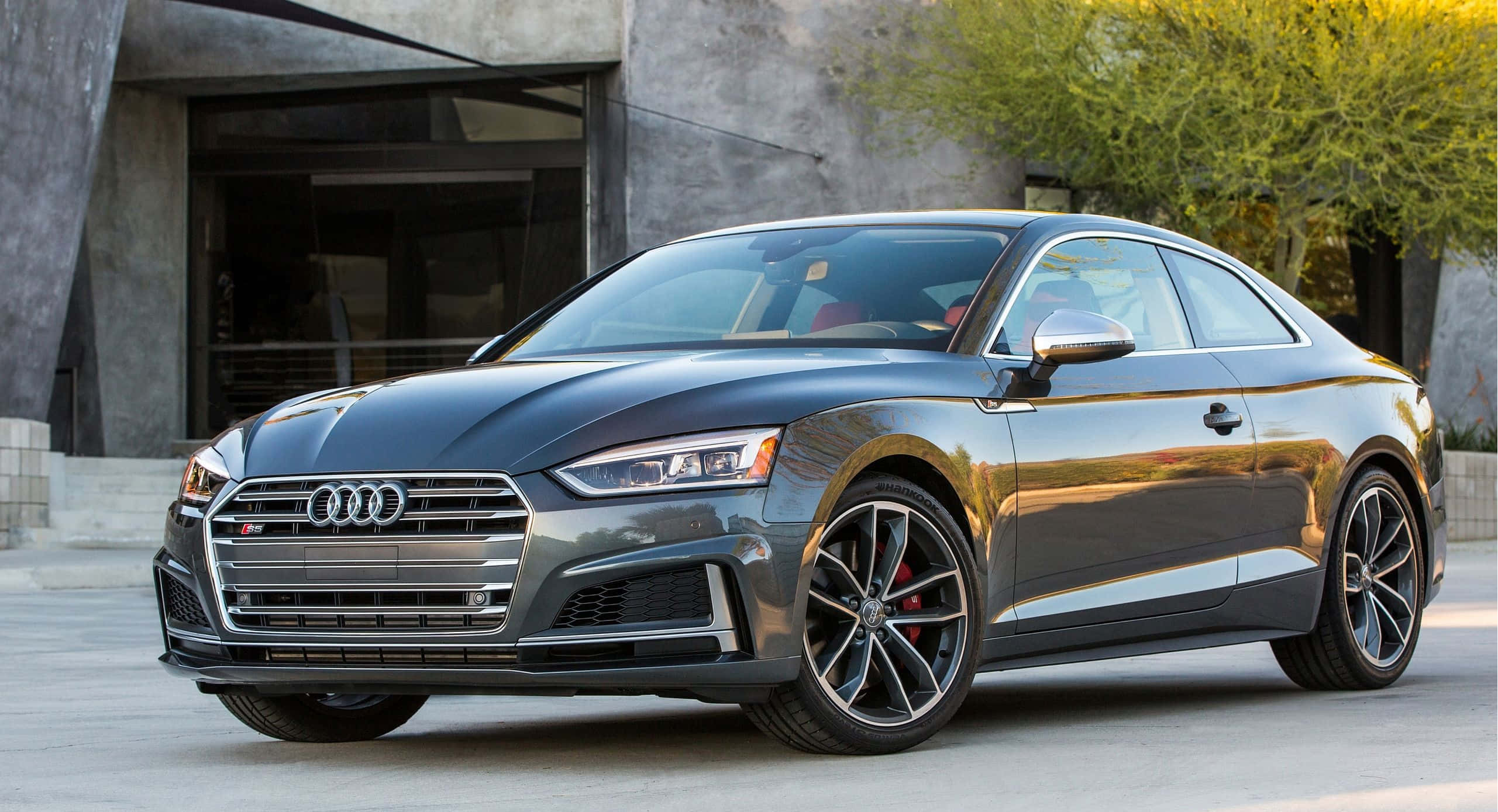 Audi S5 桌布 壁紙 — 一輛時尚的灰色奧迪S5轎跑車在金色陽光下閃閃發光，停在一棟現代混凝土建築前，附近有綠樹。