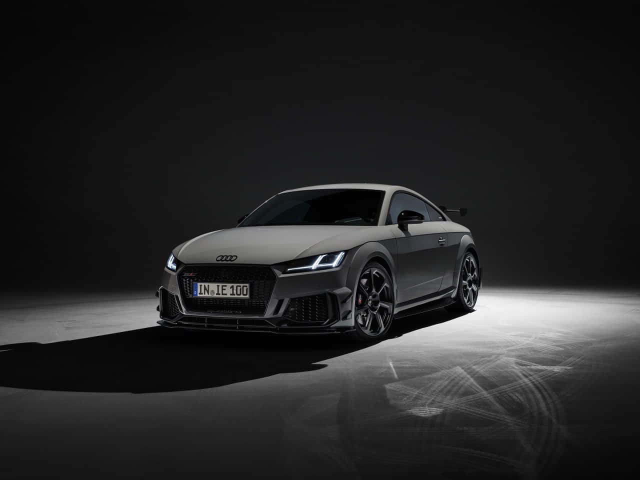 Audi Tt Rs 桌布 壁紙 — 一輛時尚的灰色奧迪TT RS雙門跑車，在黑暗的攝影棚地板上被聚光燈照亮，帶有輪胎痕跡。