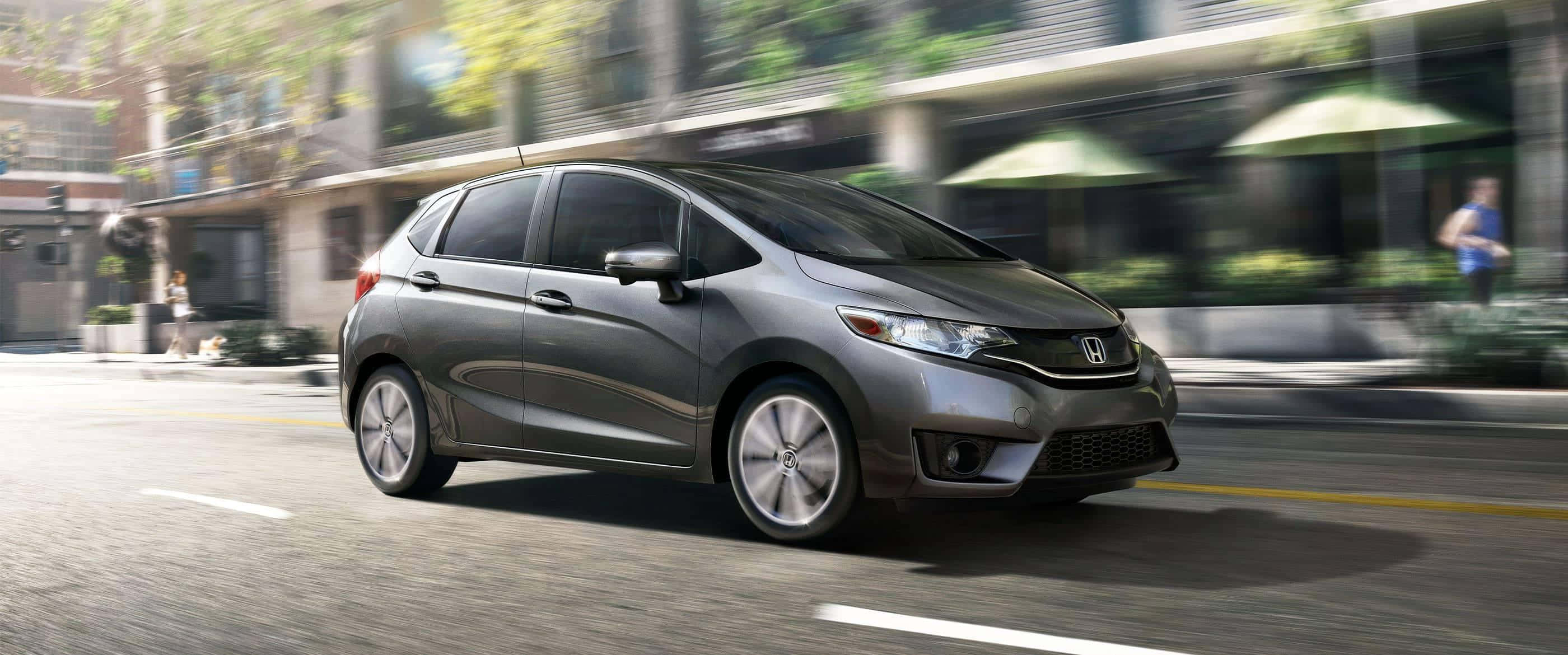 Honda Fit 桌布 壁紙 — 一輛時尚的灰色本田飛度在陽光明媚的城市街道上飛馳，背景建築物和綠色遮陽篷呈現動態模糊效果。