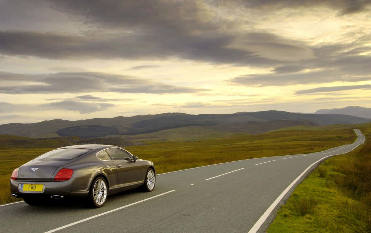 Bentley Continental Gt 桌布 壁紙 — 一輛時尚的灰色賓利雙門跑車沿著蜿蜒的鄉村道路行駛，穿過起伏的綠色山丘，天空戲劇性地多雲。