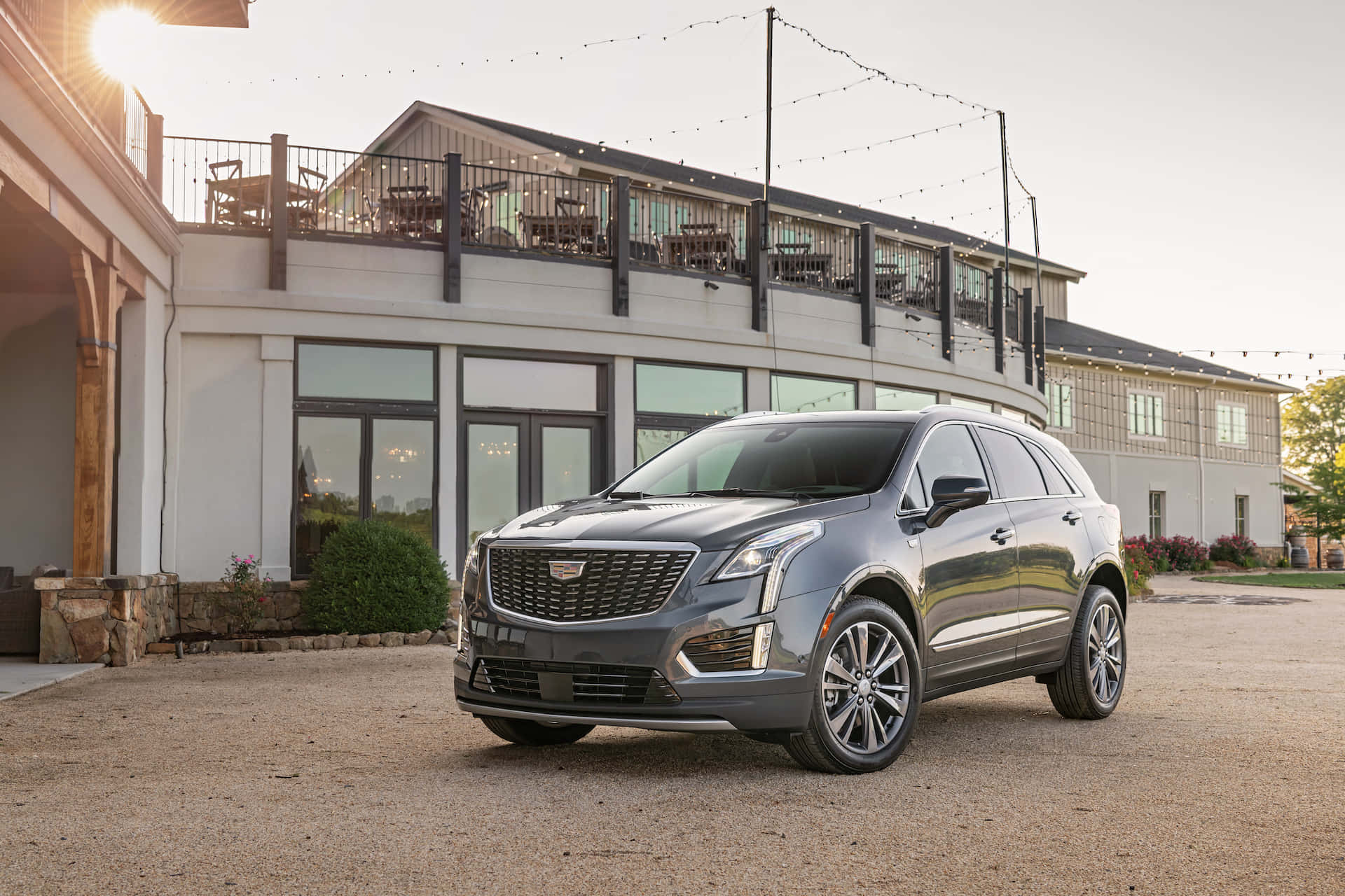 Cadillac Xt5 桌布 壁紙 — 一輛時尚的灰色 Cadillac SUV 停泊在碎石路上，前方是一座設有屋頂露台的現代建築，沐浴在金色的夕陽光中。
