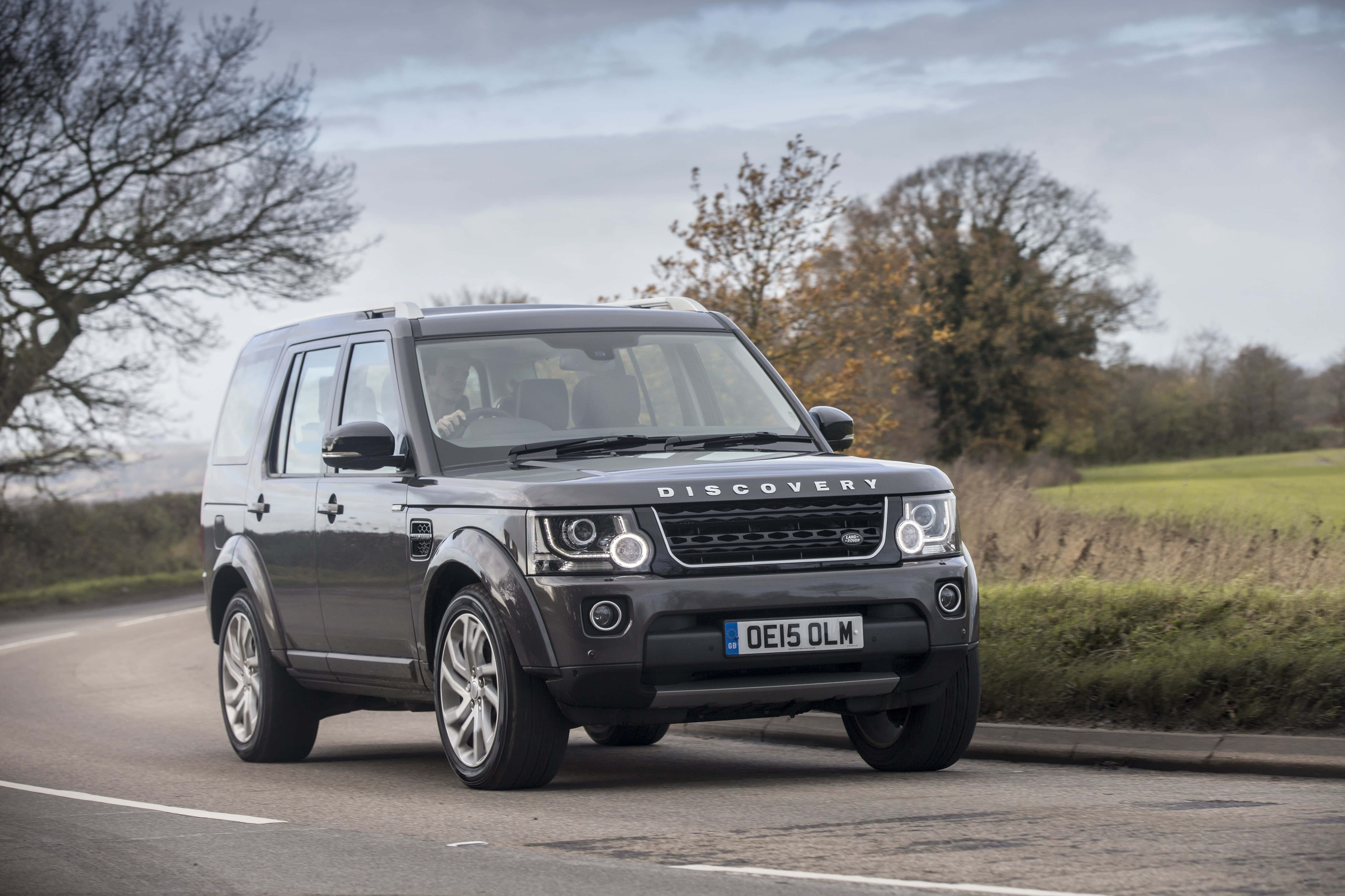 Land Rover Discovery 桌布 壁紙 — 一輛時尚的灰色Land Rover Discovery在陰天下行駛在兩旁光禿禿的樹木和秋葉的鄉村道路上。