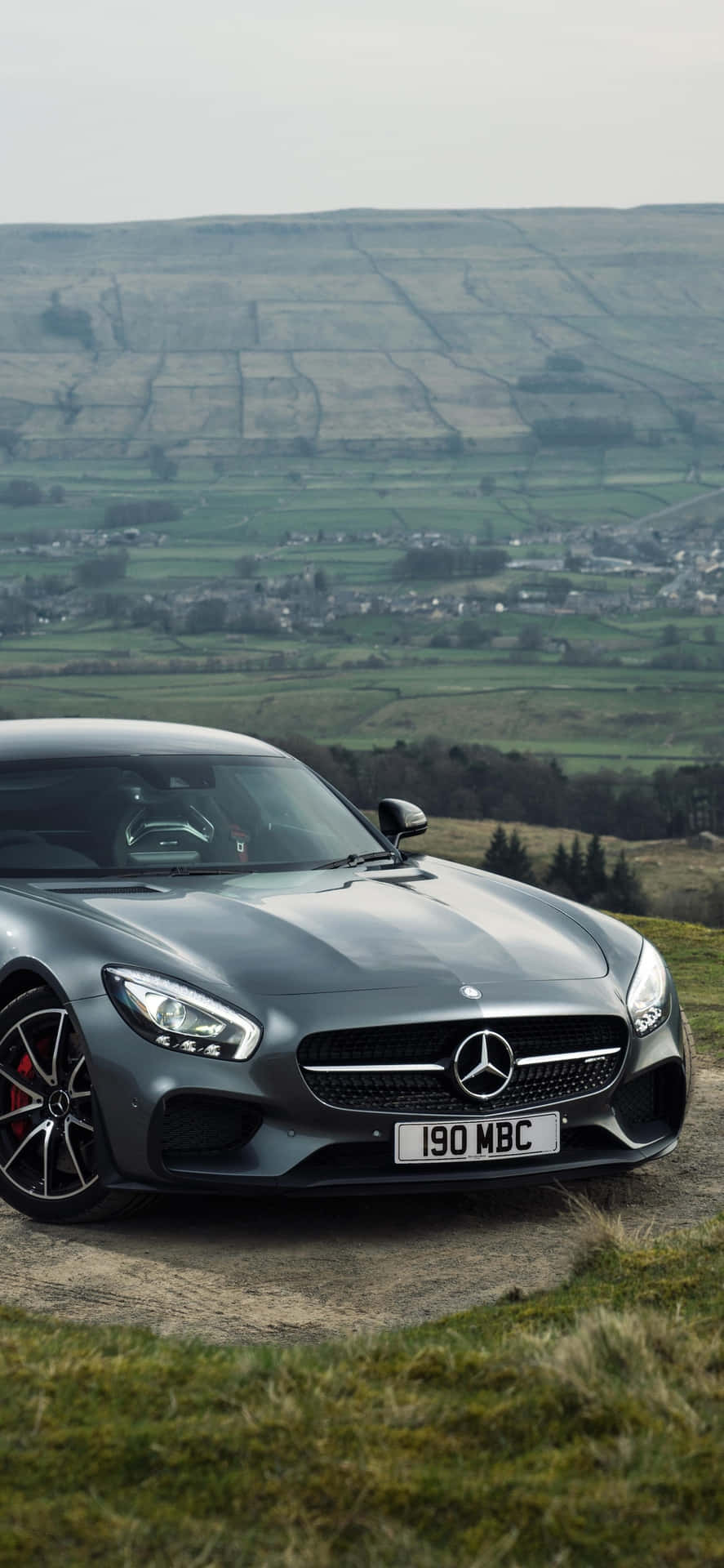 灰色Mercedes-AMG GT在一個草坡上，俯瞰著一個有拼布田野和遠處村莊的鄉村山谷。 — Amg Gt R 壁紙
