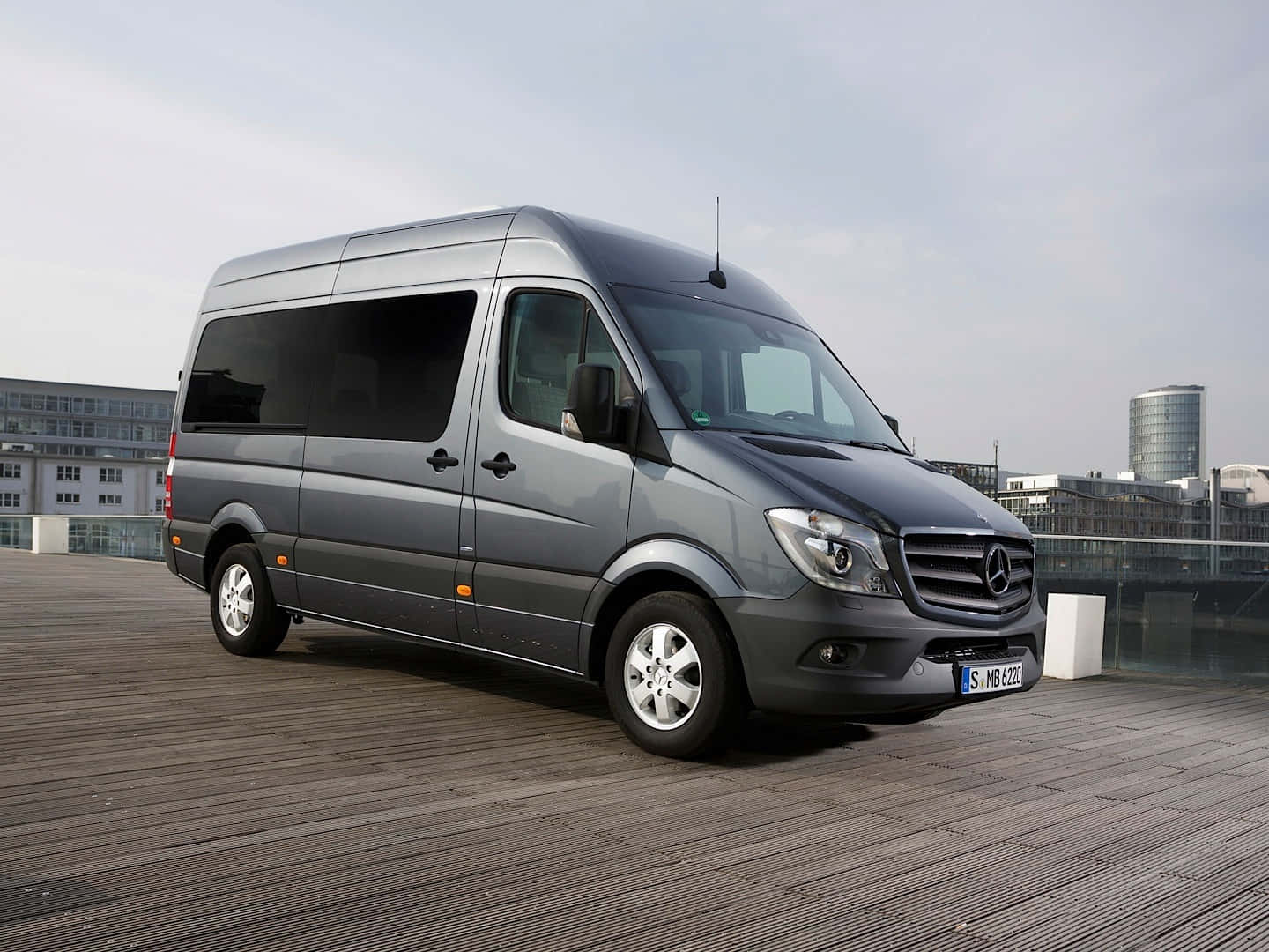 Mercedes Benz Sprinter 桌布 壁紙 — 一輛時尚的灰色 Mercedes-Benz Sprinter 客貨車停泊在木製屋頂上，背景是現代建築物，天空多雲。