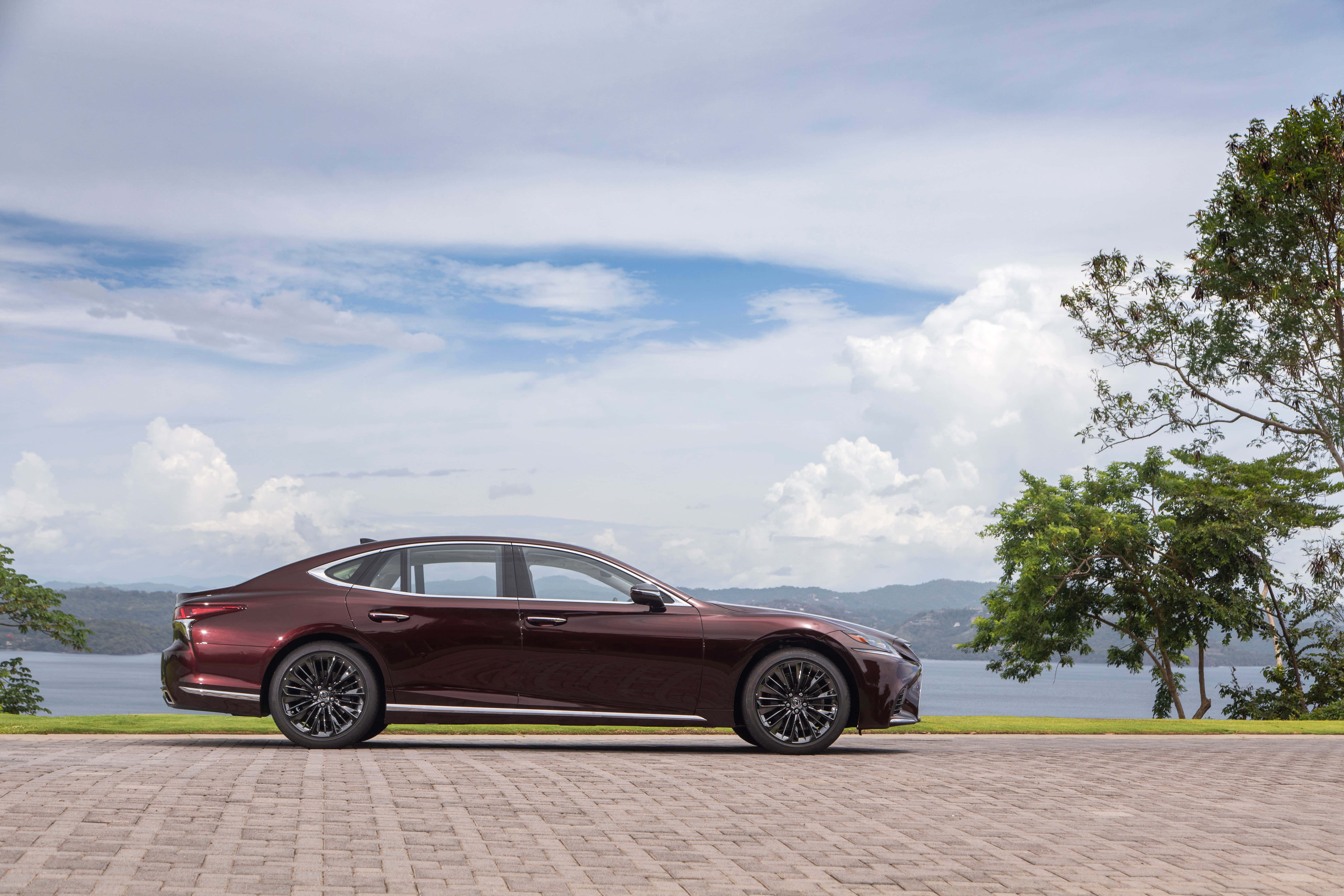Lexus Ls 500 桌布 壁紙 — 一輛時尚的酒紅色轎車停在磚砌車道上，旁邊是湖泊，遠處有山丘，頭頂有蓬鬆的雲朵。