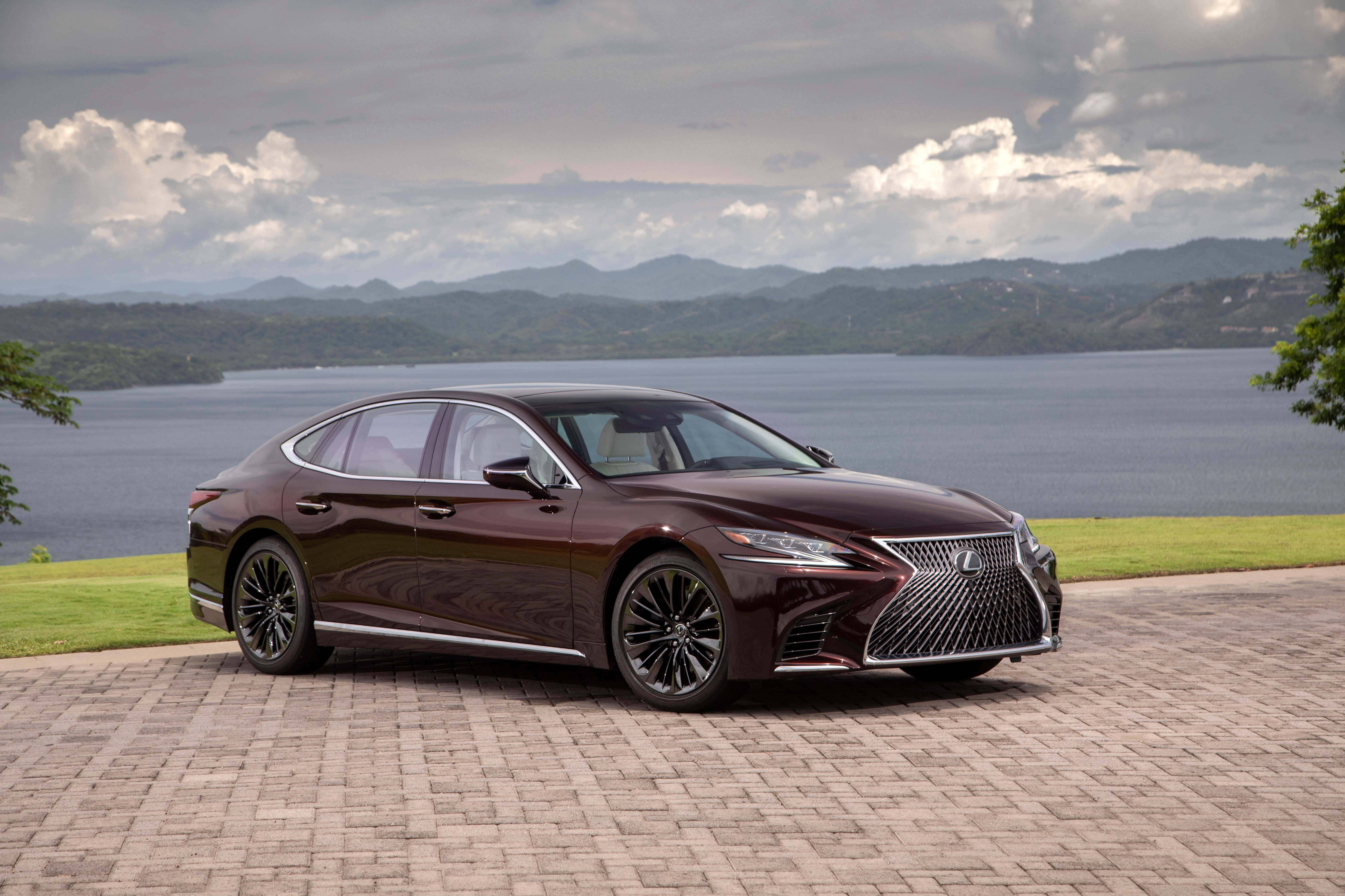 Lexus Ls 500 桌布 壁紙 — 一輛時尚的酒紅色凌志轎車停在石頭車道上，俯瞰廣闊的湖泊，遠處有起伏的山丘和陰天。