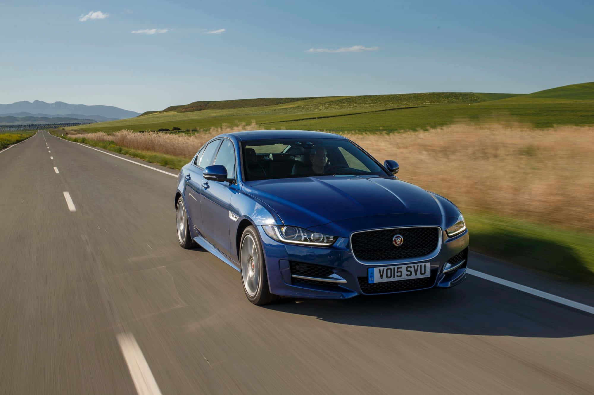 Jaguar Xe 桌布 壁紙 — 一輛時尚的藍色積架XF轎車在陽光明媚的鄉村高速公路上奔馳，沿途有連綿起伏的綠色山丘和遠處的山脈，天空清澈湛藍。