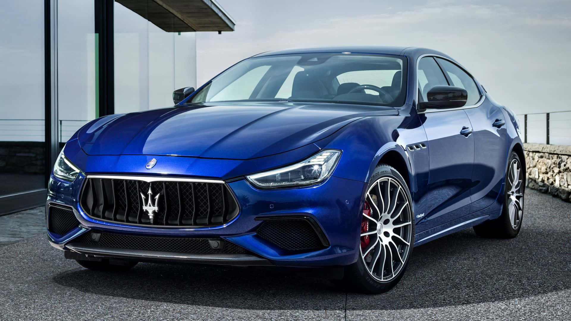 Maserati Ghibli 桌布 壁紙 — 一輛時尚的藍色瑪莎拉蒂 Ghibli 跑車停放在玻璃建築旁的現代石頭庭院上。