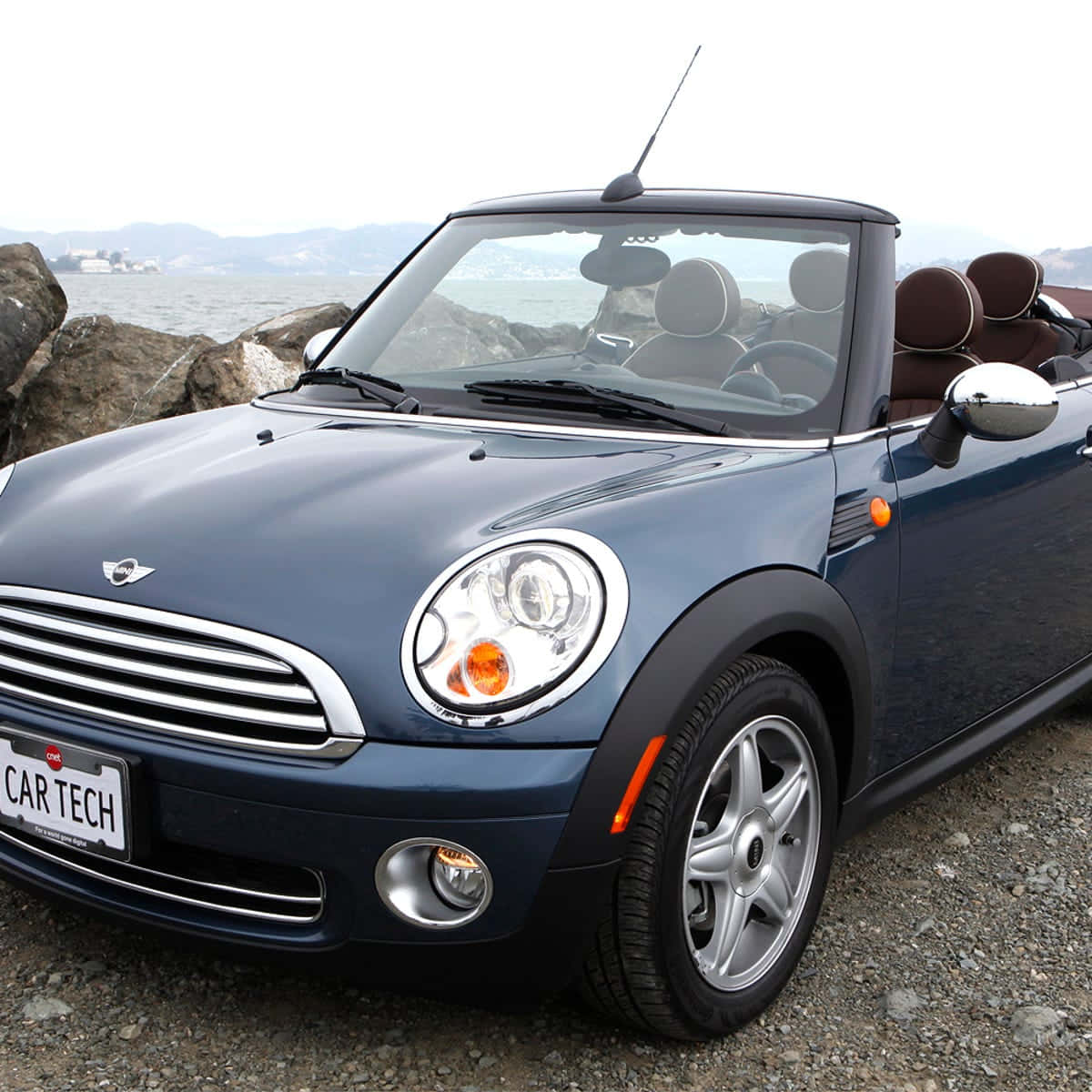 Mini Cooper Convertible 桌布 壁紙 — 一輛時尚的藍色MINI Cooper敞篷車停泊在岩石海岸上，遠處有海岸景觀，天空多雲。