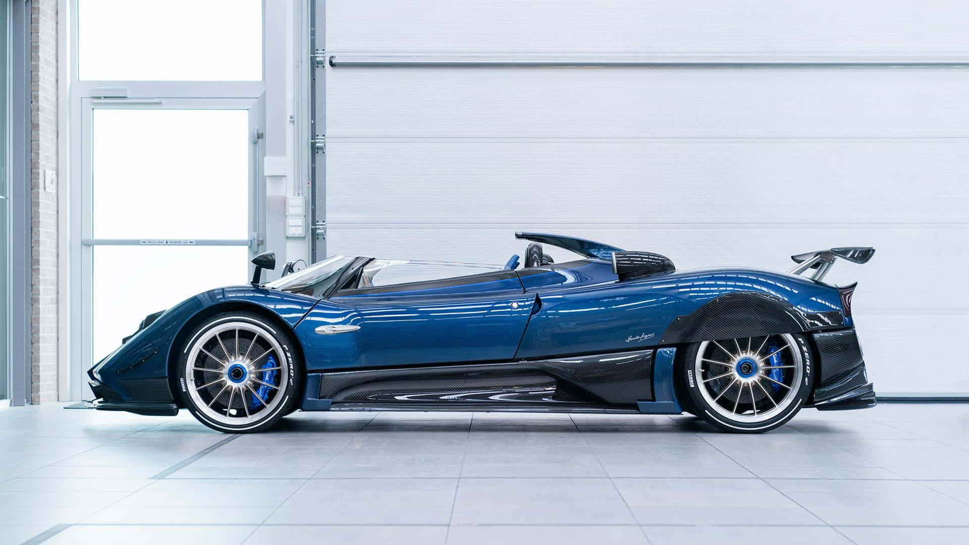 Pagani Zonda Hp Barchetta 桌布 壁紙 — 一輛時尚的藍色Pagani Zonda跑車，帶有黑色飾邊和銀色輪圈，停放在明亮的白色車庫中。