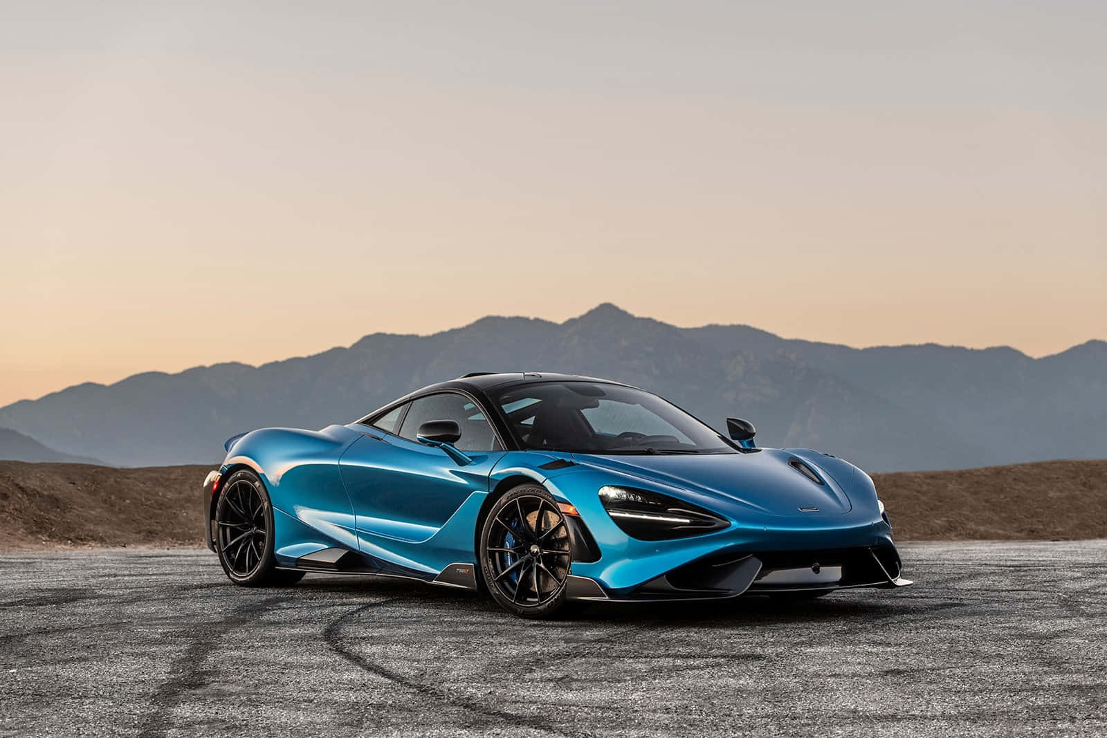 Mclaren 765lt 桌布 壁紙 — 一輛時尚的藍色跑車帶有黑色點綴，停在鋪設的賽道上，背景是日落時分的山脈。