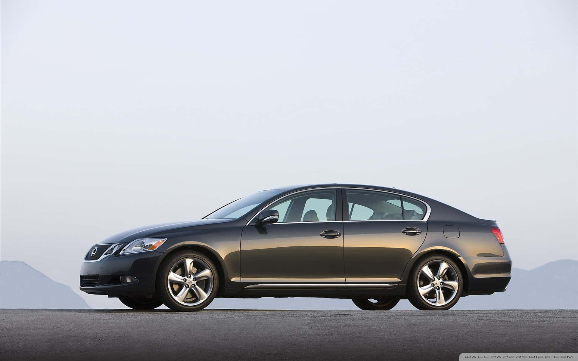 Lexus Gs 桌布 壁紙 — 一輛時尚的深灰色轎車停泊在鋪砌的道路上，遠處有山脈，天空朦朧。