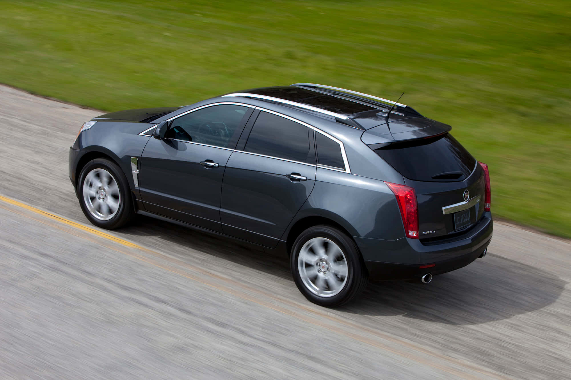 Cadillac Srx 桌布 壁紙 — 一輛時尚的深灰色凱迪拉克SRX在鋪砌的道路上行駛，背景是模糊的綠色草地。