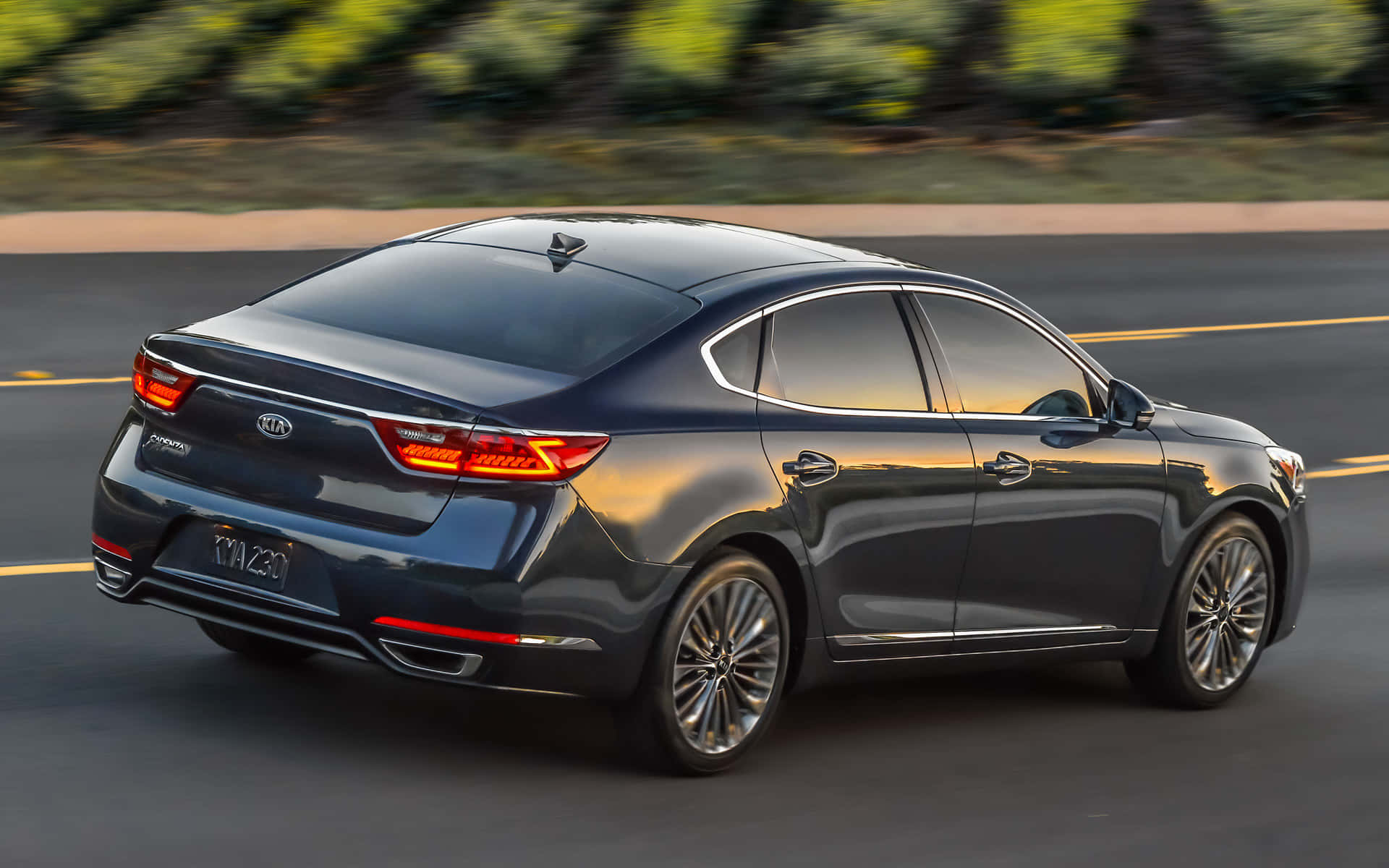 Kia Cadenza 桌布 壁紙 — 一輛時尚的深灰色Kia Optima轎車在陽光照耀的道路上平穩行駛，背景是模糊的綠色植物。