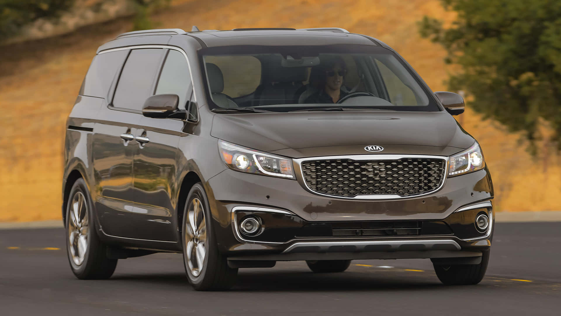 Kia Sedona 桌布 壁紙 — 一輛時尚的深灰色Kia小型貨車在道路上行駛，背景是金色的山坡和綠樹。