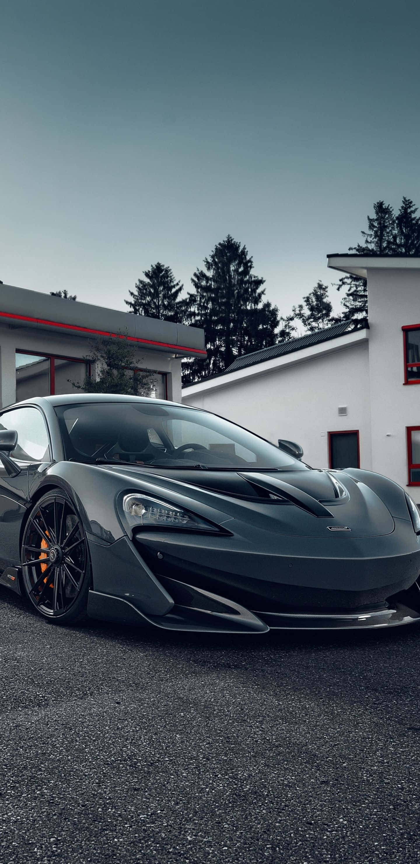 Mclaren 600lt 桌布 壁紙 — 一輛時尚的深灰色McLaren跑車停泊在瀝青路上，前方是帶有紅色點綴的現代白色建築，天空陰沉多雲。