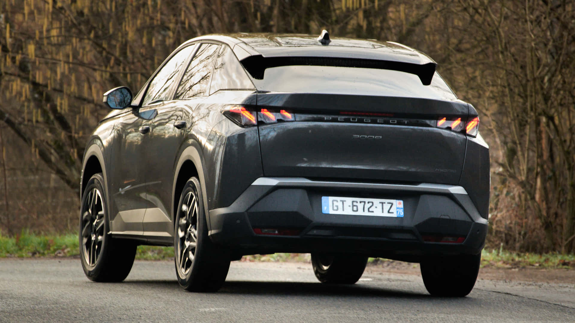  壁紙 — 一輛時尚的深灰色Peugeot 3008 SUV在秋天沿著樹木成蔭的道路行駛，周圍是光禿的樹枝和柔和的大地色調。