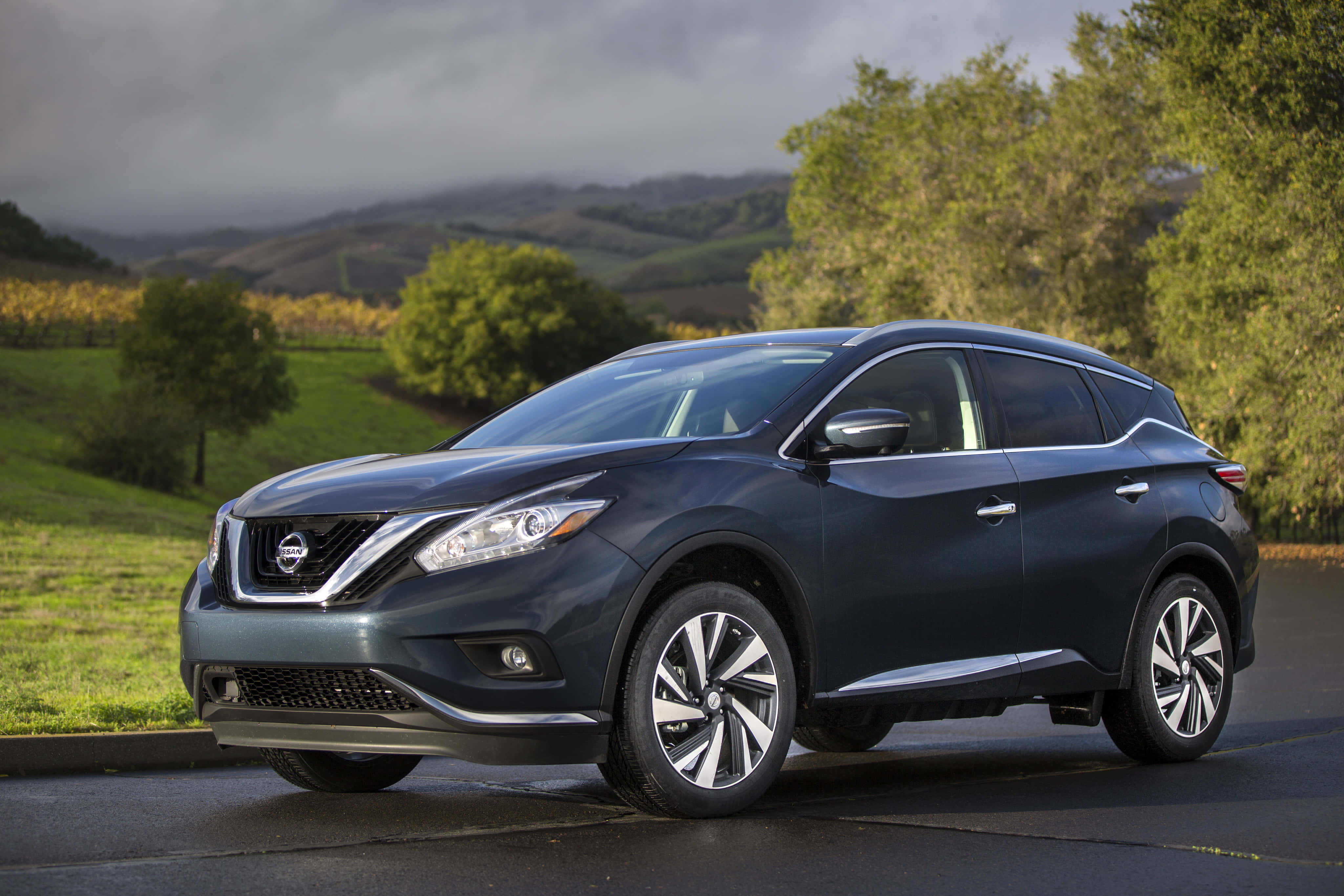 Nissan Murano 桌布 壁紙 — 一輛時尚的深藍色日產Murano SUV停泊在濕滑的道路上，背景是起伏的綠色山丘和樹木，天空多雲。