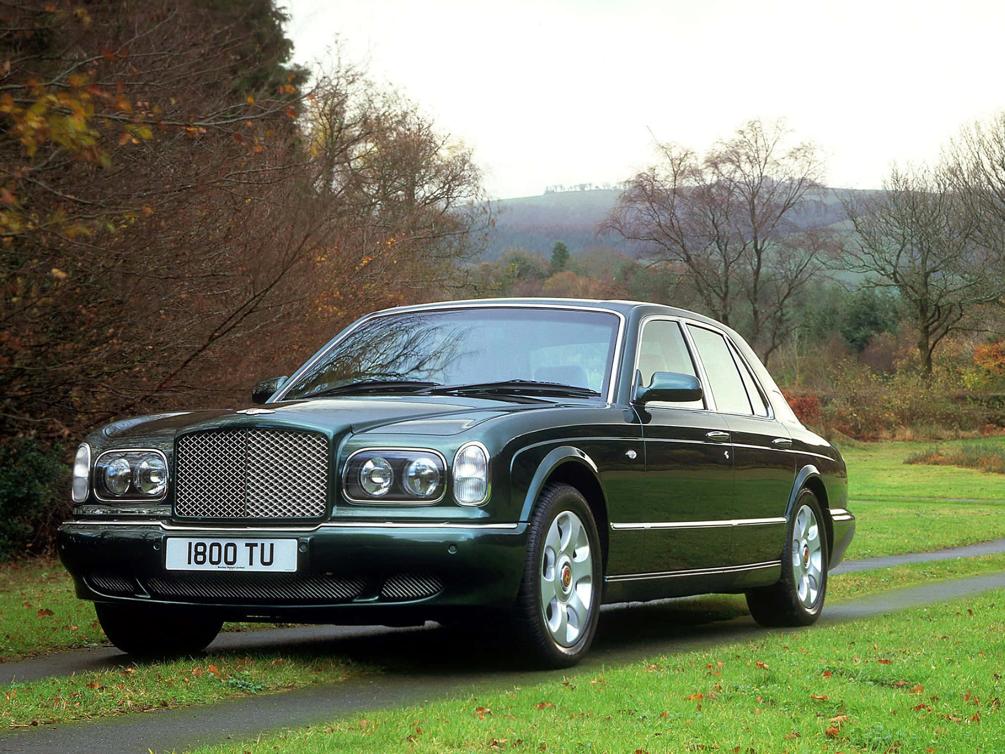 Bentley Arnage 桌布 壁紙 — 一輛時尚的深綠色賓利 Arnage 停放在蜿蜒的道路上，周圍環繞著秋天的樹木和起伏的山丘。