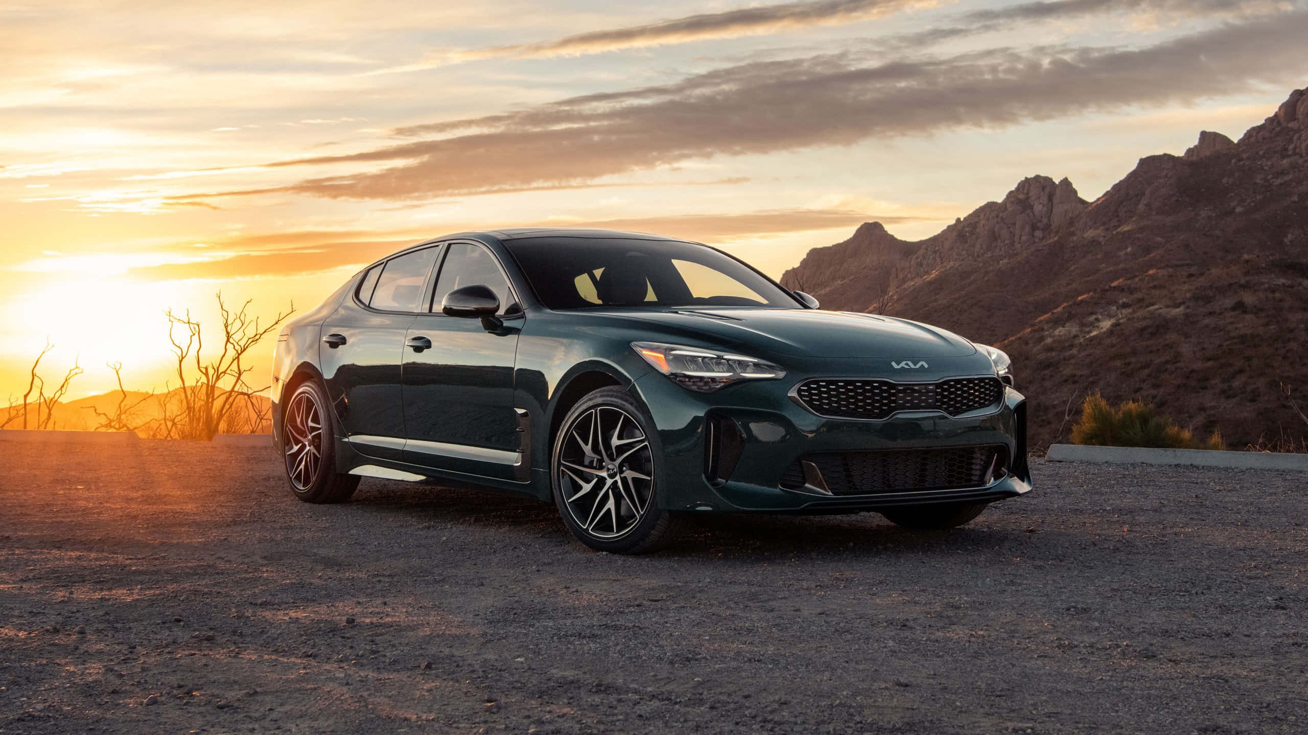 Kia Stinger 桌布 壁紙 — 一輛時尚的深綠色Kia轎車停泊在日落時分的碎石路上，背景有山脈和光禿的樹木。