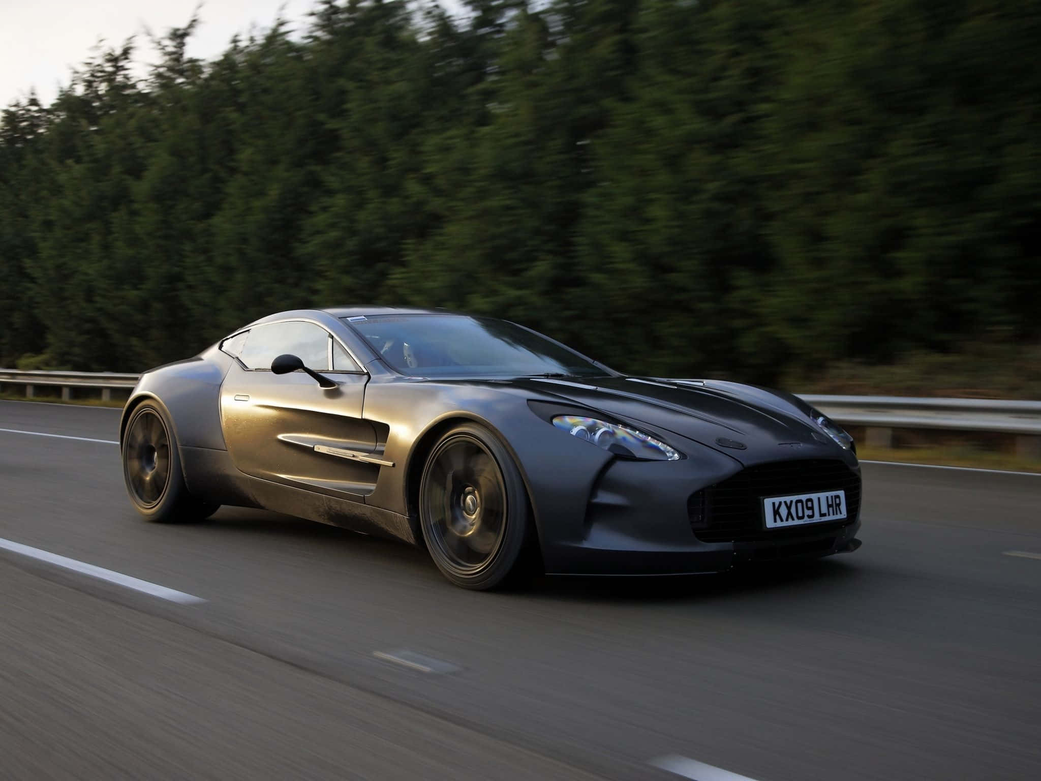 Aston Martin One-77 桌布 壁紙 — 一輛時尚的消光灰色阿斯頓馬丁One-77在森林道路上行駛，動態模糊強調了其力量和優雅。
