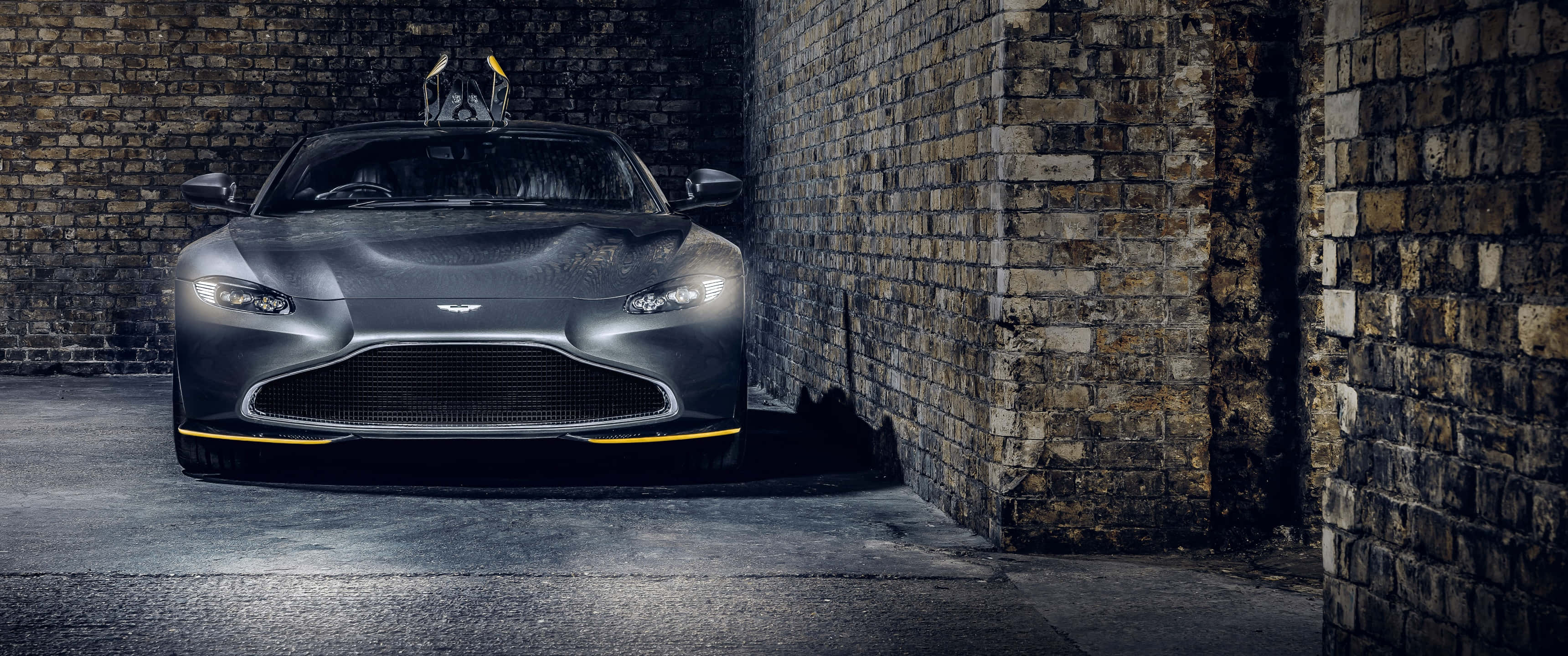 Aston Martin Vantage 桌布 壁紙 — 一輛時尚的消光灰色阿斯頓馬丁跑車，帶有黃色點綴，停在風化的磚牆之間，光線昏暗。