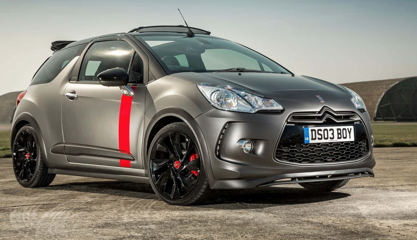  壁紙 — 一輛時尚的消光灰色 DS 3 Racing 汽車，帶有醒目的紅色條紋和黑色輪圈，停在明亮的日光下的混凝土停車場上。