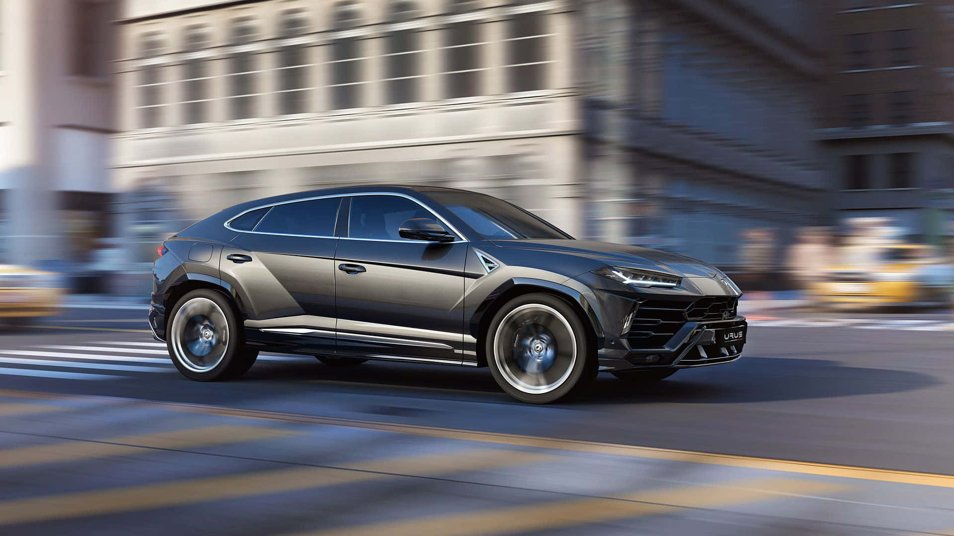 Lamborghini Urus 桌布 壁紙 — 一輛時尚的消光灰色藍寶堅尼 Urus 在模糊的城市街道上奔馳，動態模糊強調了它的力量和速度。