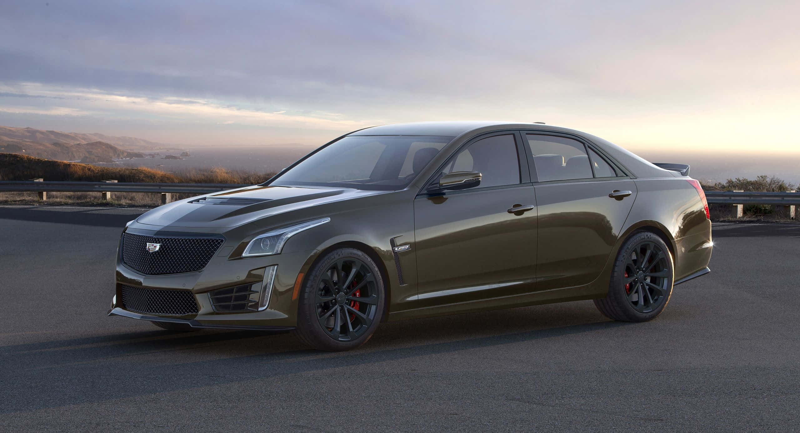 Cadillac Cts 桌布 壁紙 — 一輛時尚的啞光橄欖綠色凱迪拉克CTS-V轎車，配備黑色輪圈和紅色制動卡鉗，停在日落時分的沿海公路上。