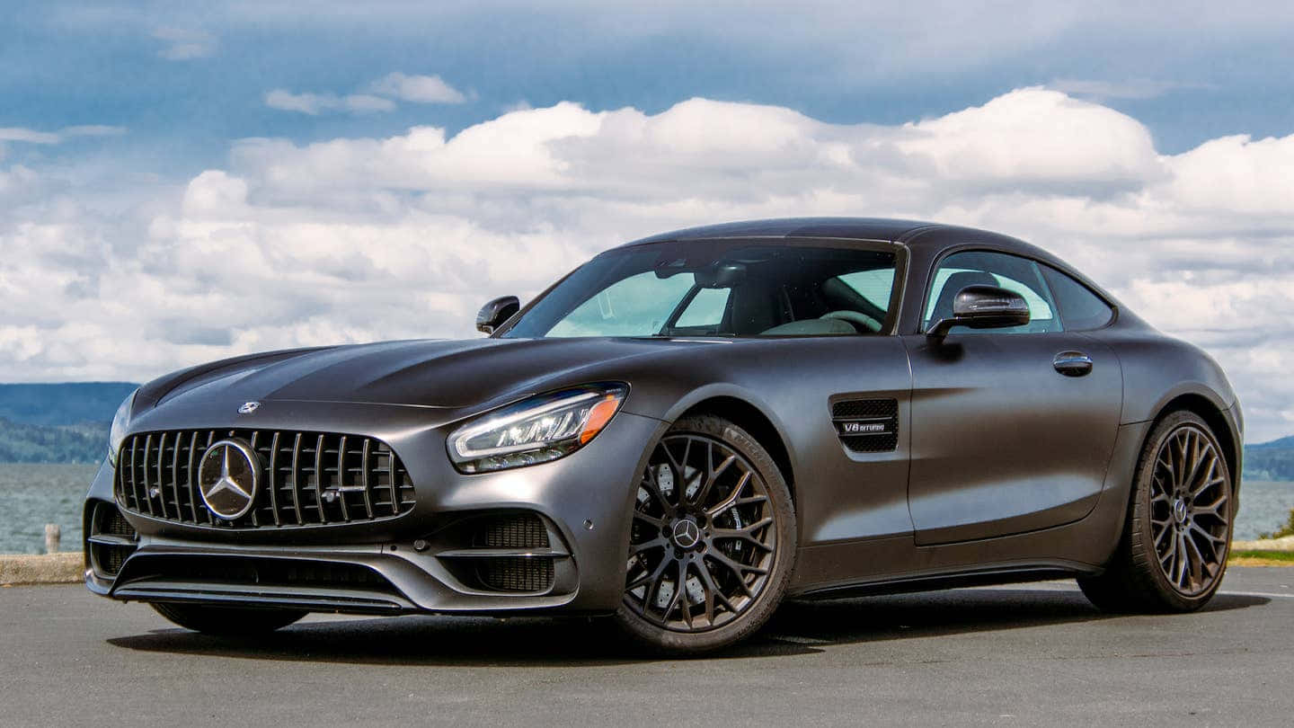 Mercedes Benz Amg Gt 桌布 壁紙 — 一輛時尚的啞光灰色梅賽德斯-AMG GT跑車停泊在雲層密佈的天空下的沿海道路上。