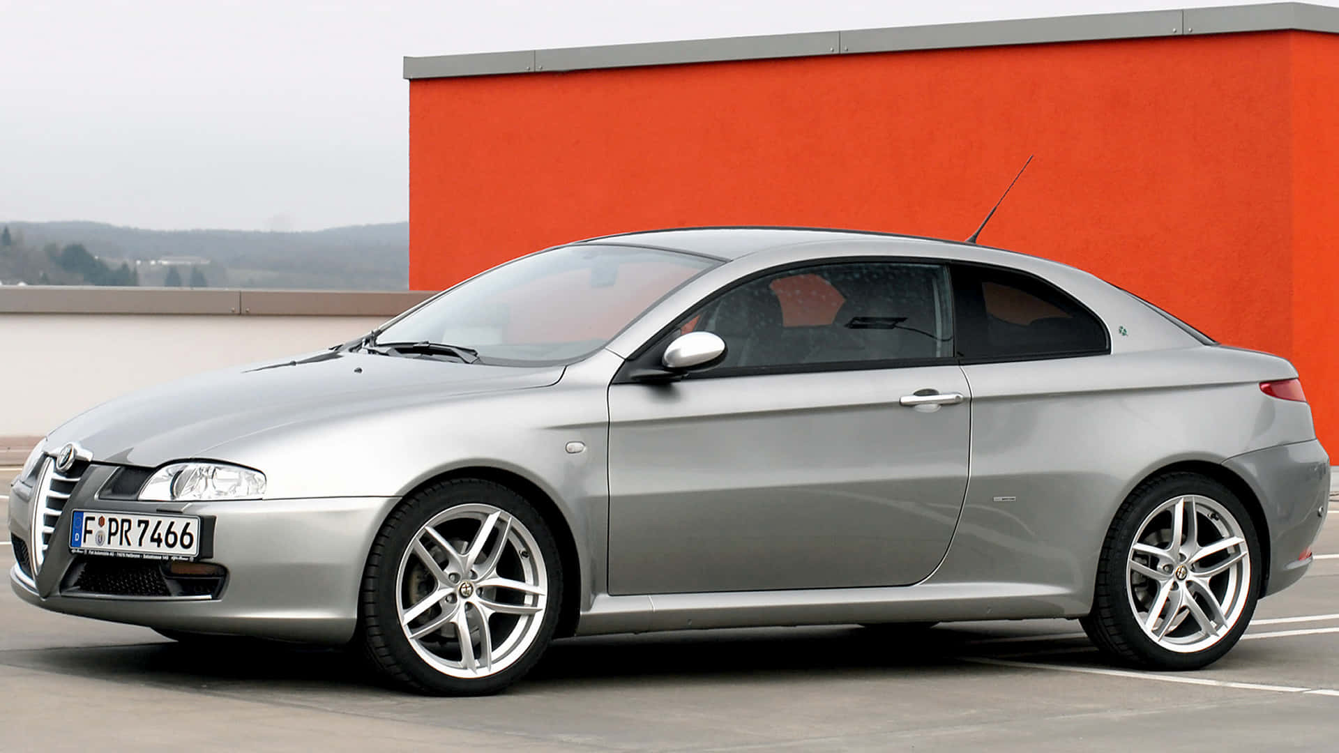 Alfa Romeo Gt 桌布 壁紙 — 一輛時尚的銀色愛快羅密歐雙門跑車停泊在陰天下一道大膽的橙色牆旁邊。