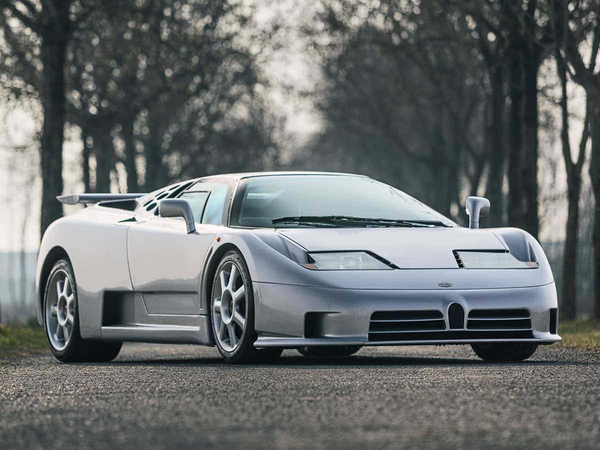 Bugatti Eb110 桌布 壁紙 — 一輛時尚的銀色Bugatti EB110超級跑車停泊在碎石路上，兩旁是光禿禿的樹木，在柔和日光下。