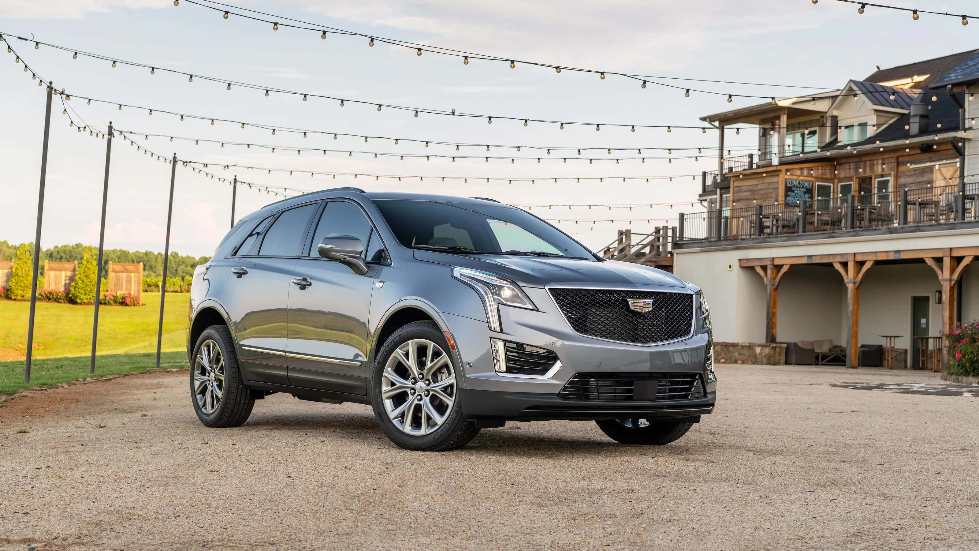 Cadillac Xt5 桌布 壁紙 — 一輛時尚的銀色Cadillac SUV停泊在碎石路上，前面是一間帶有串燈的質樸木屋。