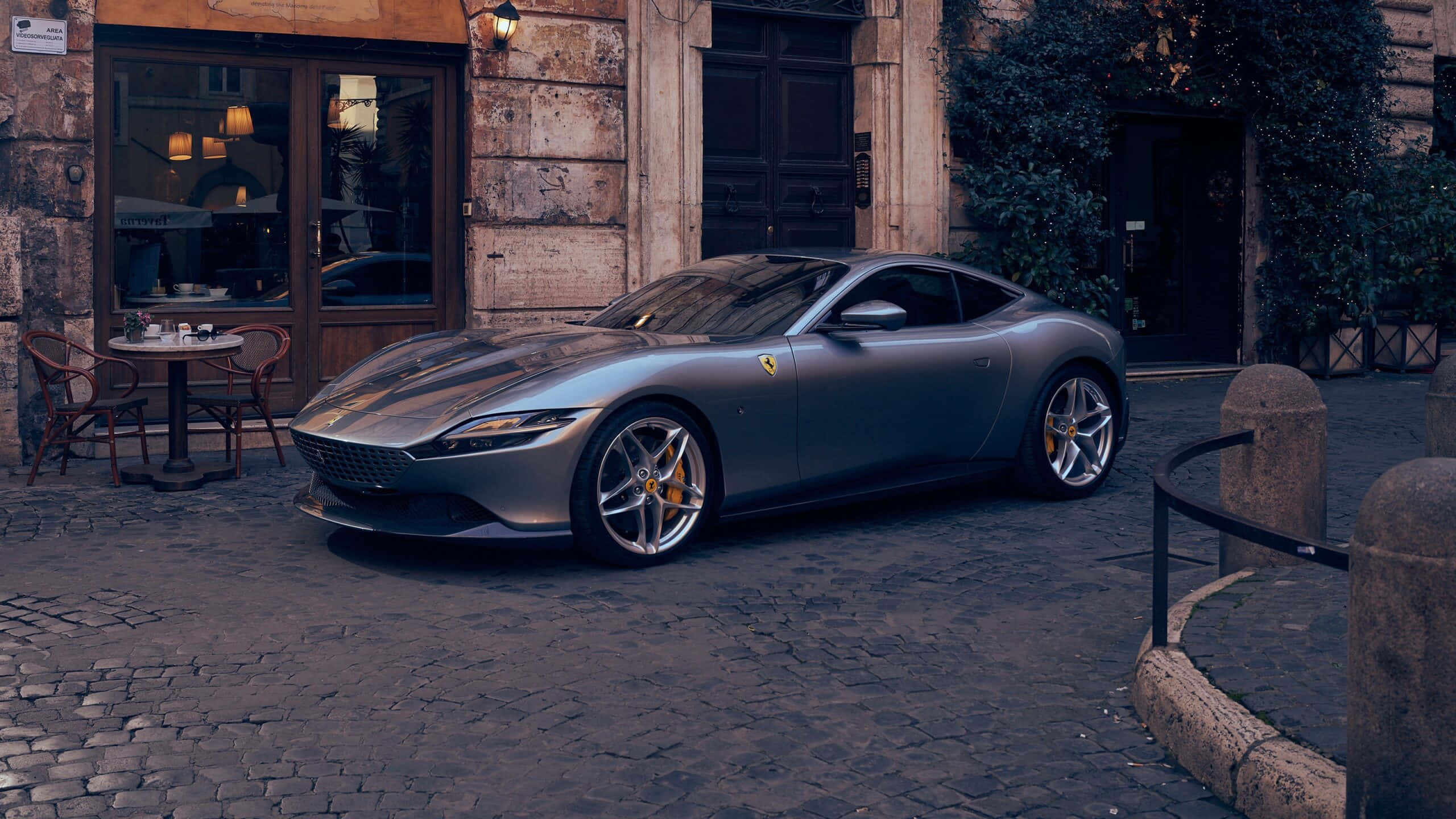 Ferrari Roma 桌布 壁紙 — 一輛時尚的銀色法拉利跑車停泊在鵝卵石街道上，旁邊是一間帶有溫暖室內燈光的鄉村風格咖啡館。