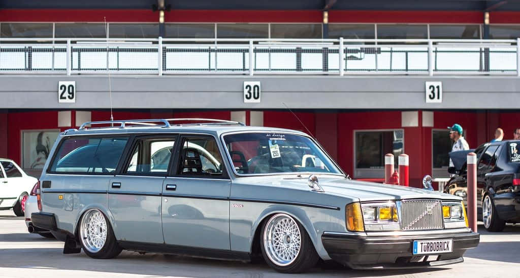Volvo Pv544 桌布 壁紙 — 一輛時尚的銀色富豪240旅行車，配備定制輪圈，在明亮的日光下停泊在賽車場的維修區。