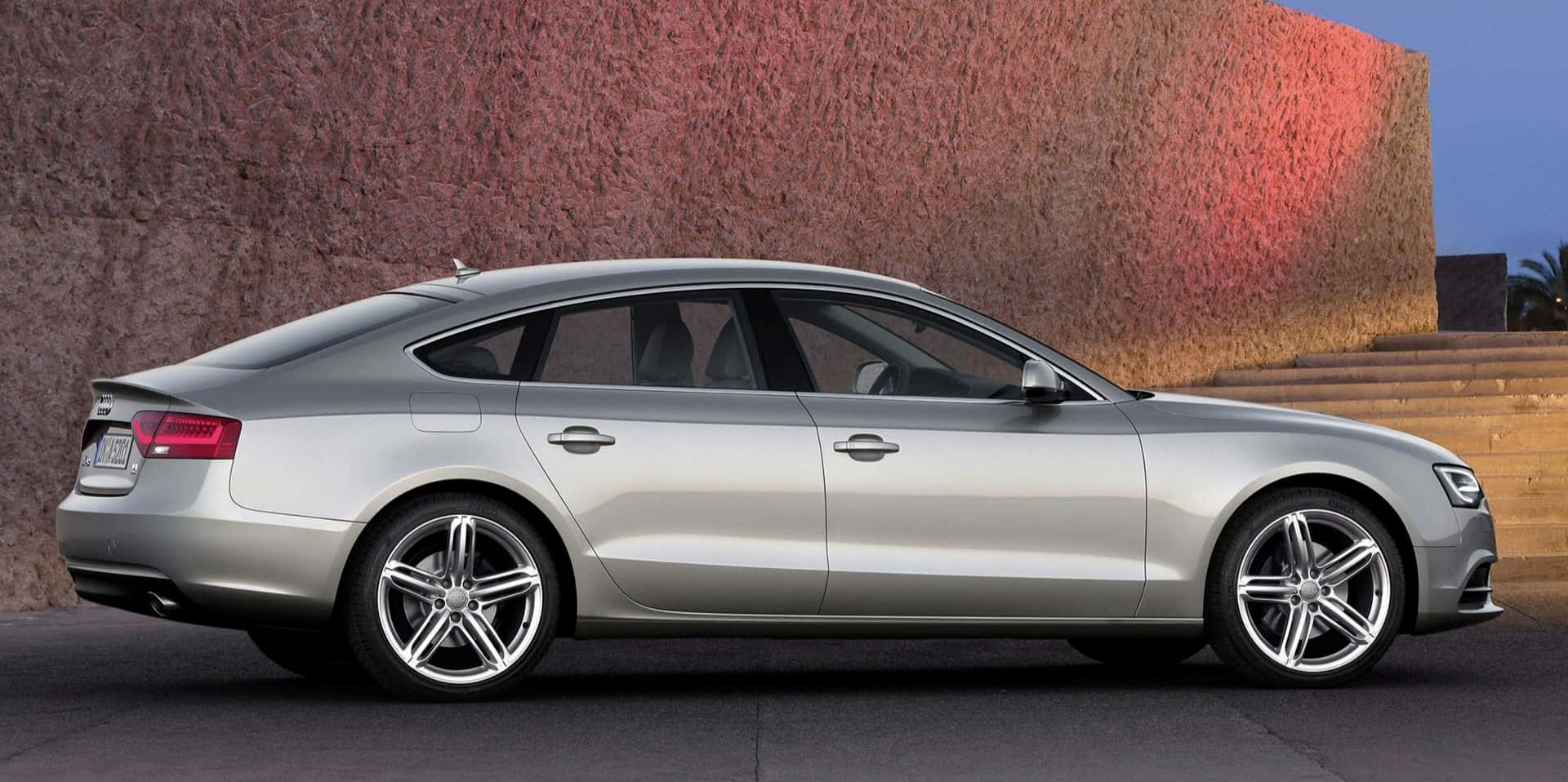 Audi A5 桌布 壁紙 — 一輛時尚的銀色轎車停泊在暮色燈光下帶紋理的微紅色牆壁旁。
