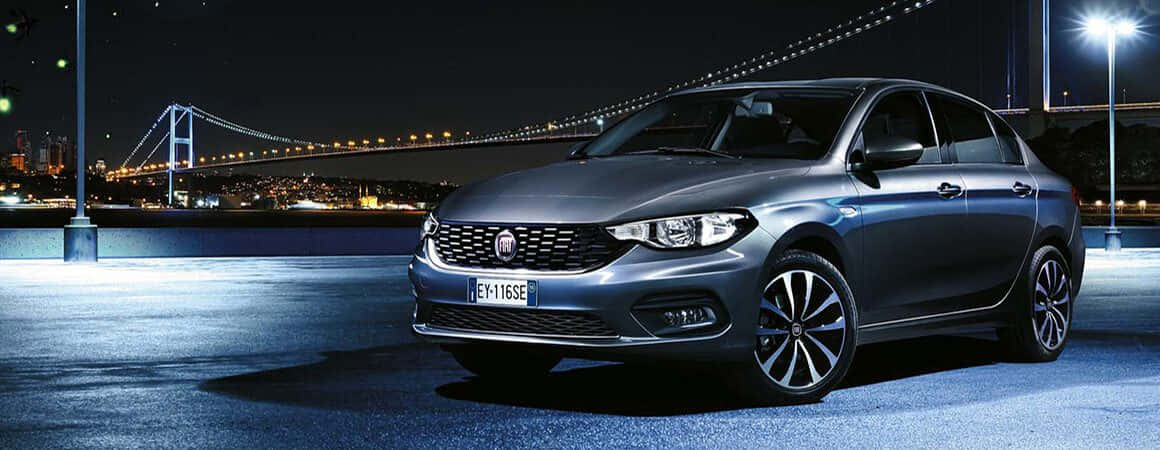 Fiat Tipo 桌布 壁紙 — 一輛時尚的銀色轎車在夜間停泊在明亮的燈光下，背景是發光的橋樑和城市景觀。