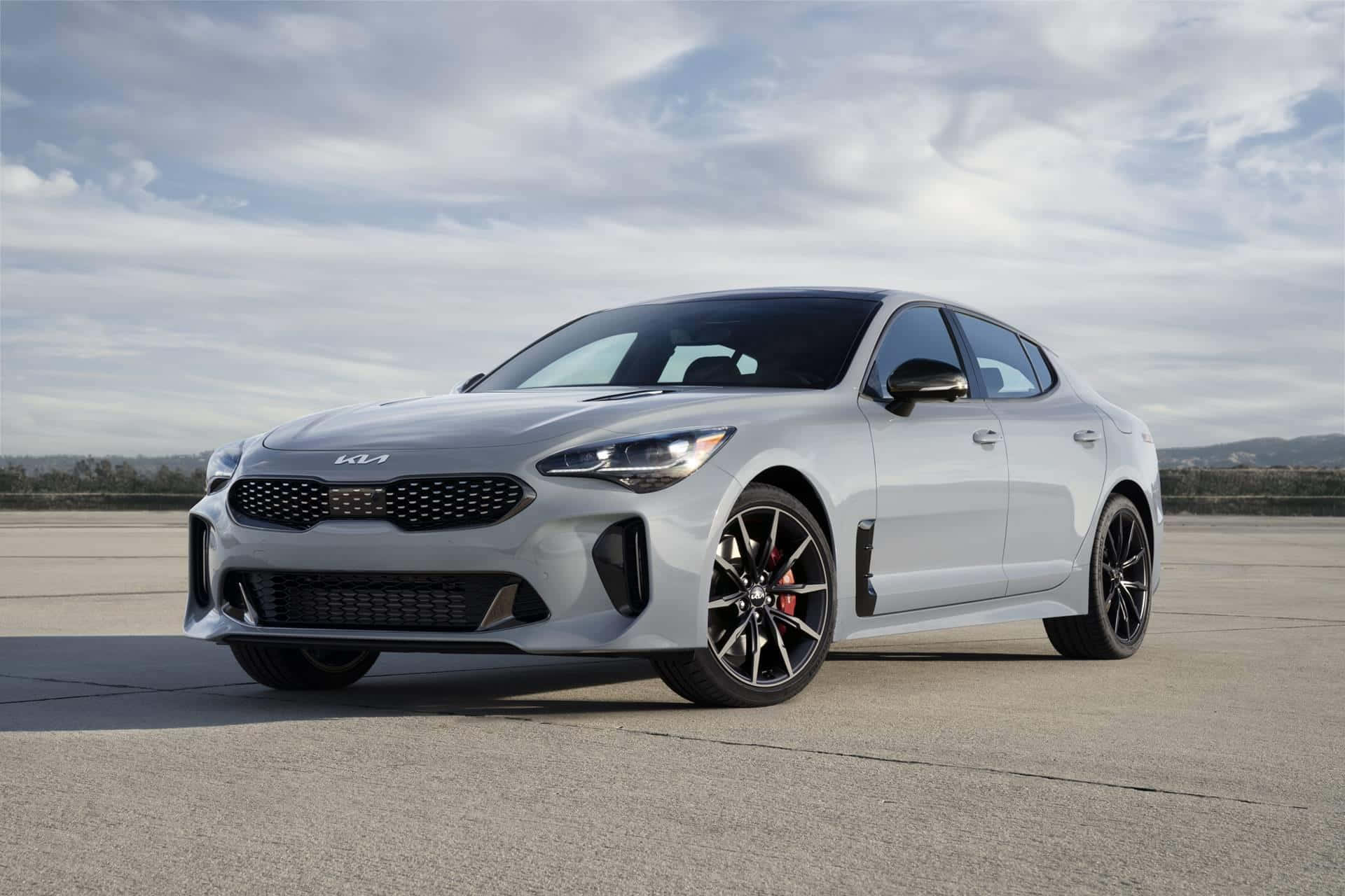 Kia Stinger 桌布 壁紙 — 一輛時尚的銀色Kia Stinger運動轎車，帶有黑色飾件和紅色制動卡鉗，停放在混凝土停車場上，天空多雲。
