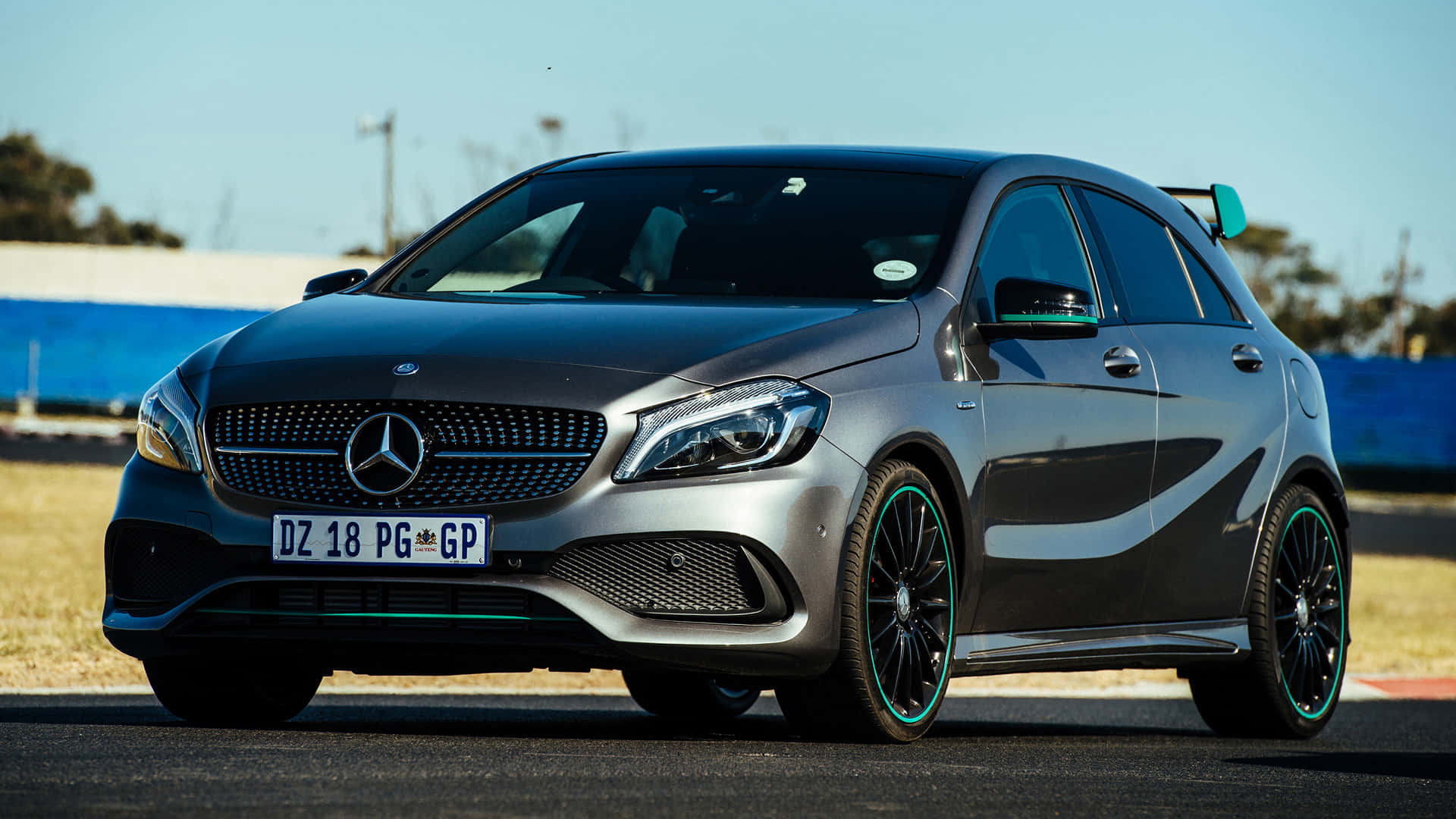 Mercedes Benz A-class 桌布 壁紙 — 一輛時尚的銀色 Mercedes-AMG A45 S 跑車，配備綠色點綴的輪圈，停在瀝青賽道上，天空湛藍。