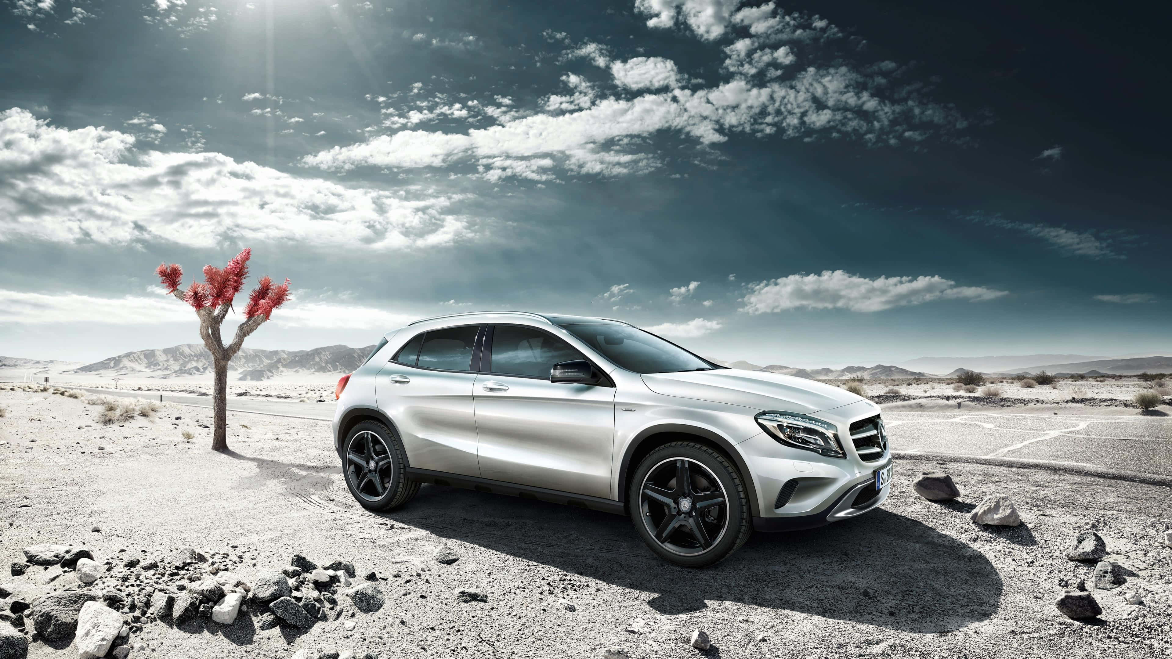 Mercedes Benz Gla-class 桌布 壁紙 — 一輛時尚的銀色SUV停泊在沙漠公路上，旁邊有一棵孤獨的紅葉樹，天空佈滿戲劇性的雲層。