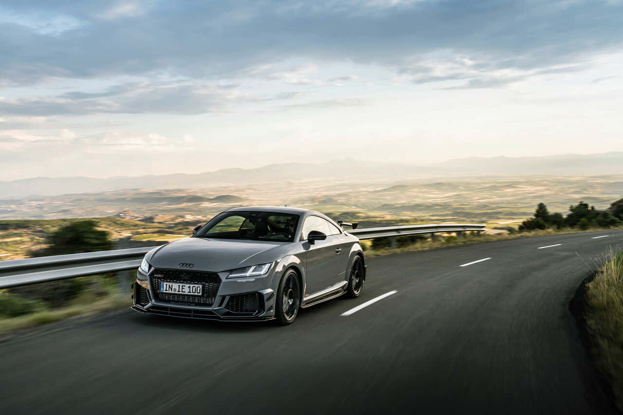Audi Tt Rs 桌布 壁紙 — 一輛時尚的銀色奧迪TT RS在雲層密布的天空和遠山背景下，沿著蜿蜒的山路飛馳。