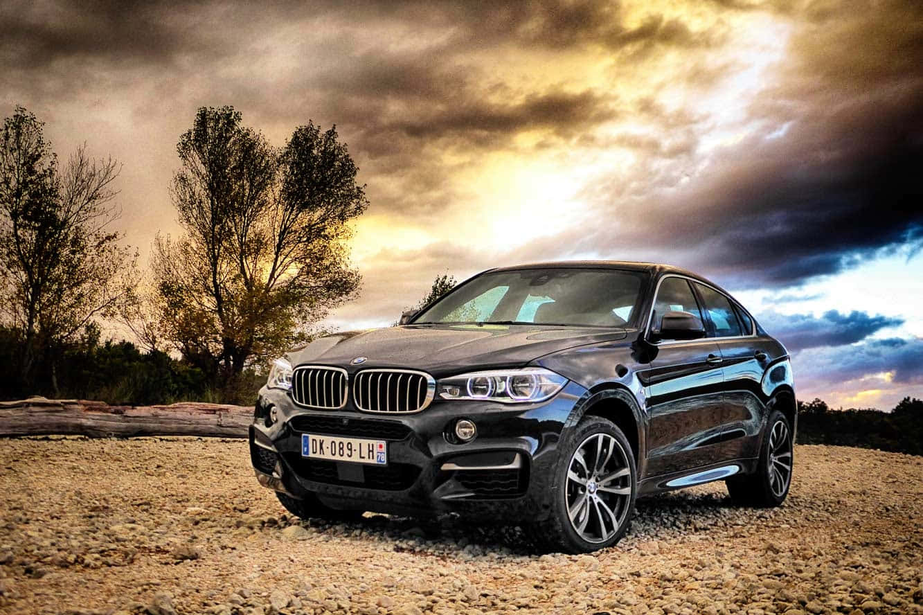 Bmw X6 桌布 壁紙 — 一輛時尚黑色BMW X6 SUV停泊在碎石場上，背景是戲劇性的日落天空，金色雲朵和樹木剪影。