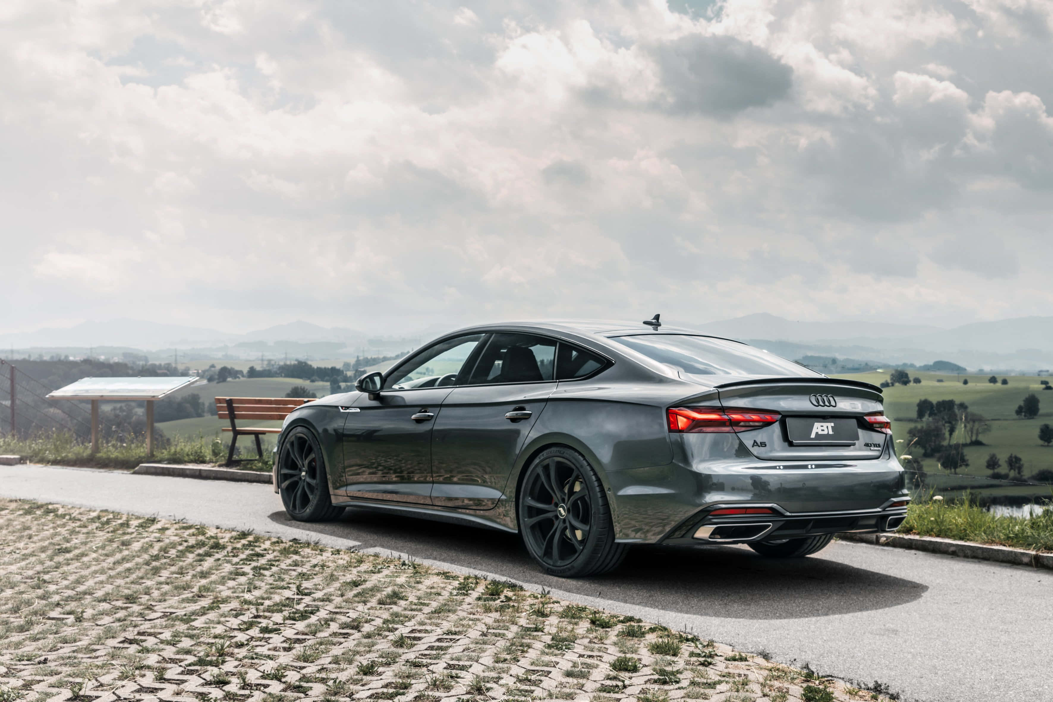 Audi A5 桌布 壁紙 — 一輛時尚灰色ABT Audi A5 Sportback停泊在鵝卵石路上，俯瞰著多雲天空下連綿起伏的綠色山丘。