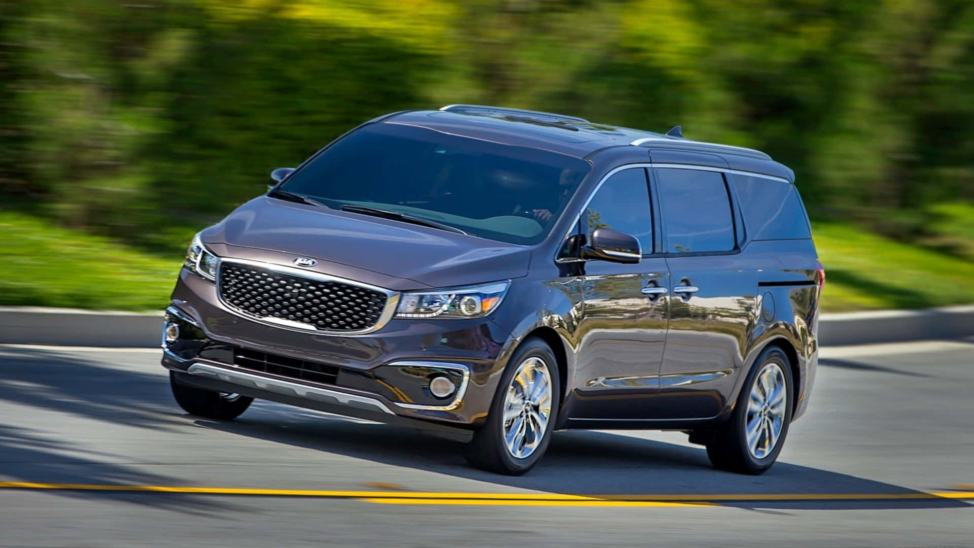 Kia Sedona 桌布 壁紙 — 一輛時尚深灰色Kia小型客車在陽光明媚的道路上行駛，背景有模糊的綠色樹葉。